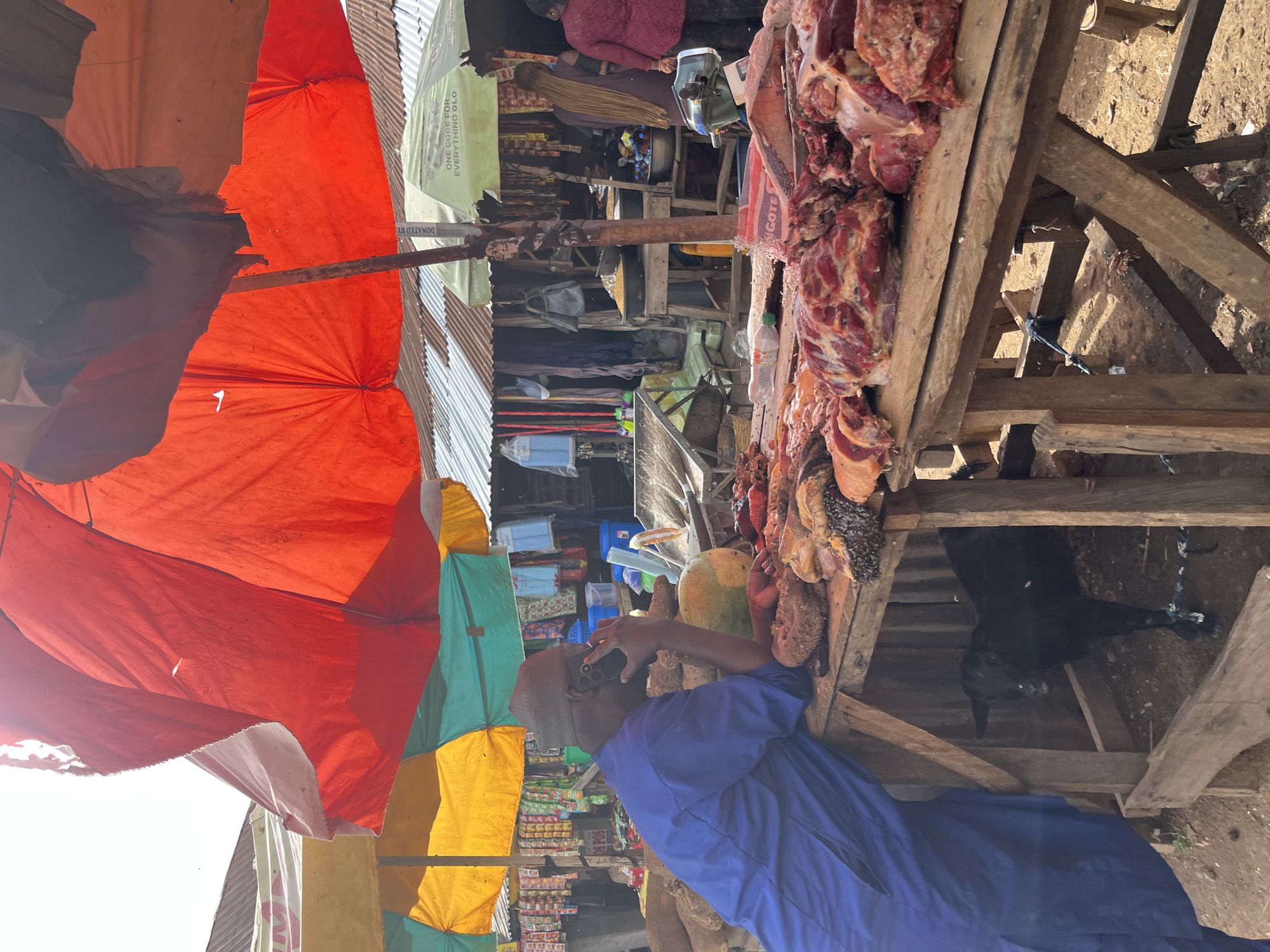 This is a forensic, matter-of-fact description of a market scene, possibly in Rafin Par, Nigeria. A man wearing a blue robe and a dark hat is standing at a wooden table laden with raw meat. He is holding a dark-colored phone to his ear, appearing to be in a conversation. The table is covered with various cuts of meat, some of which show signs of being prepared for sale. Flies are visible on the meat, indicating a typical outdoor market environment.
Behind the man and the meat stall, a small black goat is visible, tethered within a wooden enclosure. The market is covered by large, brightly colored tarpaulin umbrellas, with red, yellow, and green sections visible. In the background, stalls are set up with various goods displayed, including packaged items, clothing, and what appear to be grains or dry goods in bowls. Another person, a woman with long braided hair and wearing a brown outfit, is seen in the background, as is another woman in a red top. The overall environment suggests an active, open-air marketplace.