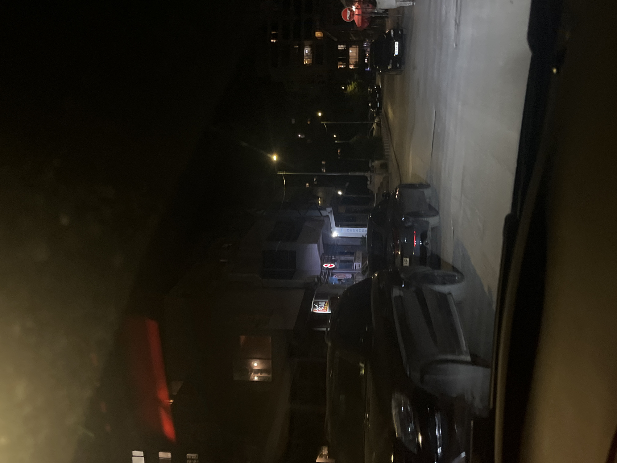Captured from the vantage point within a vehicle, the image reveals a nocturnal urban setting. The presence of streetlights illuminates the scene, casting light onto the adjacent buildings and street. A variety of vehicles populate the road, implying vehicular traffic, although no pedestrians are directly visible. Architectural details of the surrounding buildings are discernible, hinting at residential or commercial properties. Based on architectural style and street layout, the location seems to be Sofia, Bulgaria.