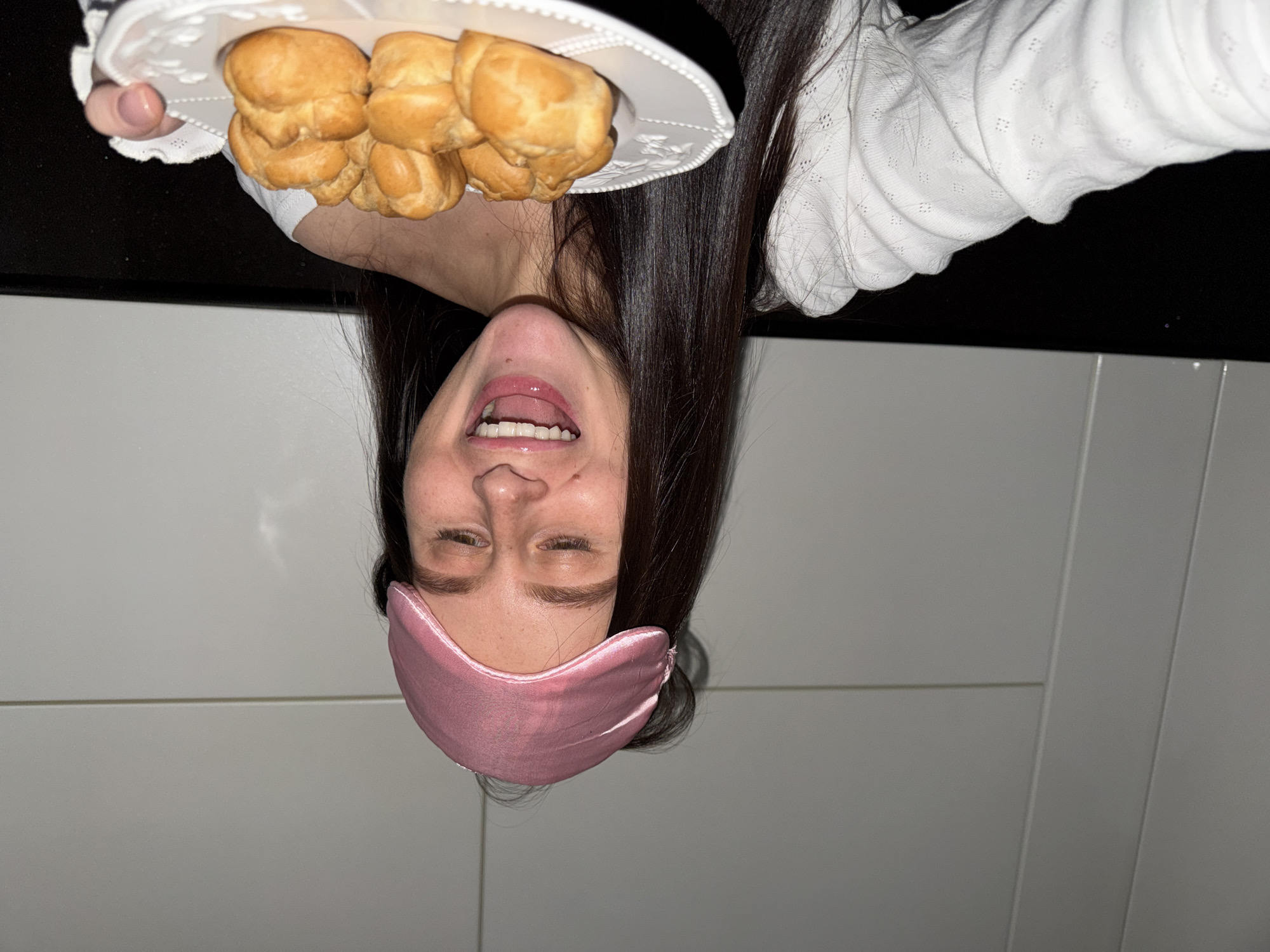 A young woman is captured in a playful and joyful moment, holding a plate of pastries above her head. The image is oriented vertically, with her head appearing at the bottom when viewed normally.
The subject is a young woman with long, dark, straight hair. She is wearing a white long-sleeved top with a subtle textured or eyelet pattern. A pink sleep mask is pushed up on her face, revealing her upper face and squinting eyes, which convey a sense of delight or amusement. Her mouth is wide open in a smile, with her tongue slightly out, suggesting anticipation or excitement.
She is holding a white decorative plate filled with several golden-brown, puffed pastries, resembling profiteroles or cream puffs. Her right thumb, visible holding the plate, has light pink nail polish.
The background is simple and domestic, consisting of a dark surface (possibly a counter or tabletop) to her upper left and light-colored, paneled walls behind her and to her right. The lighting appears to be artificial, indicating an indoor setting. The overall scene suggests a spontaneous and cheerful moment, possibly involving a late-night or early-morning snack at home in Khimki, Russia. There is no visible text in the image.