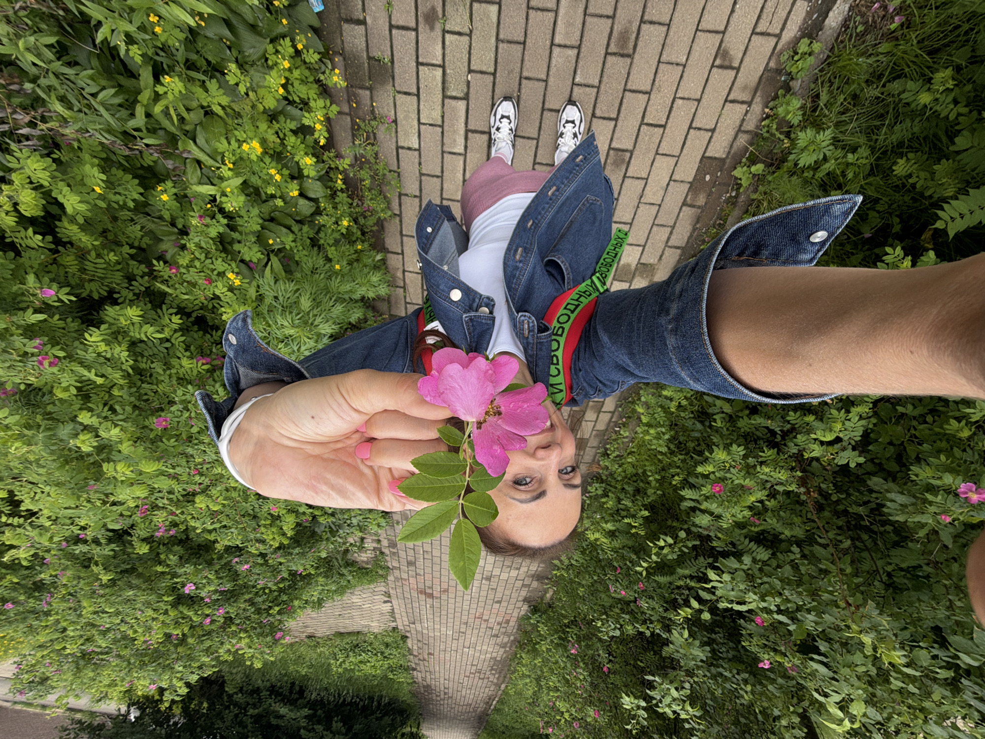 A woman is holding a pink flower with green leaves in her right hand, extending it towards the camera. She is wearing a denim jacket, a white t-shirt, pink leggings, and white sneakers with black accents. She has a red backpack with a green strap visible over her shoulders. The woman is standing on a brick pathway lined with lush green bushes and small pink and yellow flowers. The pathway recedes into the distance, suggesting an outdoor, park-like setting. The overall impression is that of a selfie taken during a walk. The location is identified as Birobidzhan, Russia.