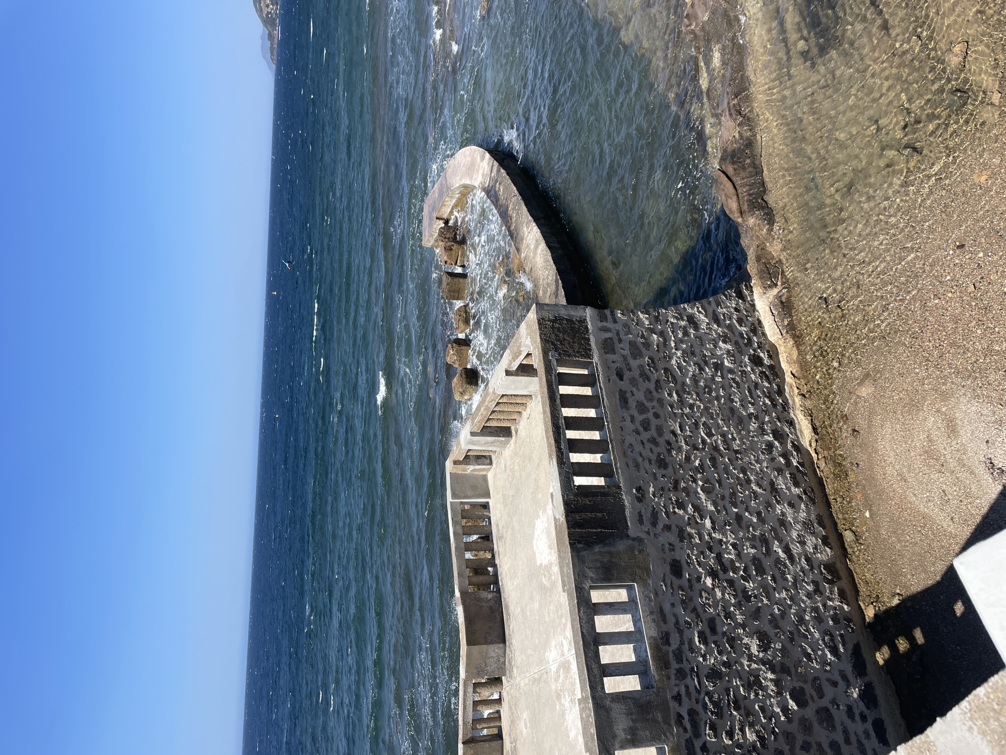 A paved viewing platform with a stone-clad base overlooks the Mediterranean Sea in Carqueiranne, France. The platform features a concrete floor and a low concrete balustrade with rectangular openings. Adjacent to the platform, a curved concrete seawall encloses a small section of the sea where rocks are visible. The water is a deep blue, with gentle waves and white foam. A sandy and pebbly shore is visible in the lower right of the image. The sky is clear and blue, with distant hazy landmasses on the horizon. There are no people or animals visible in the scene.