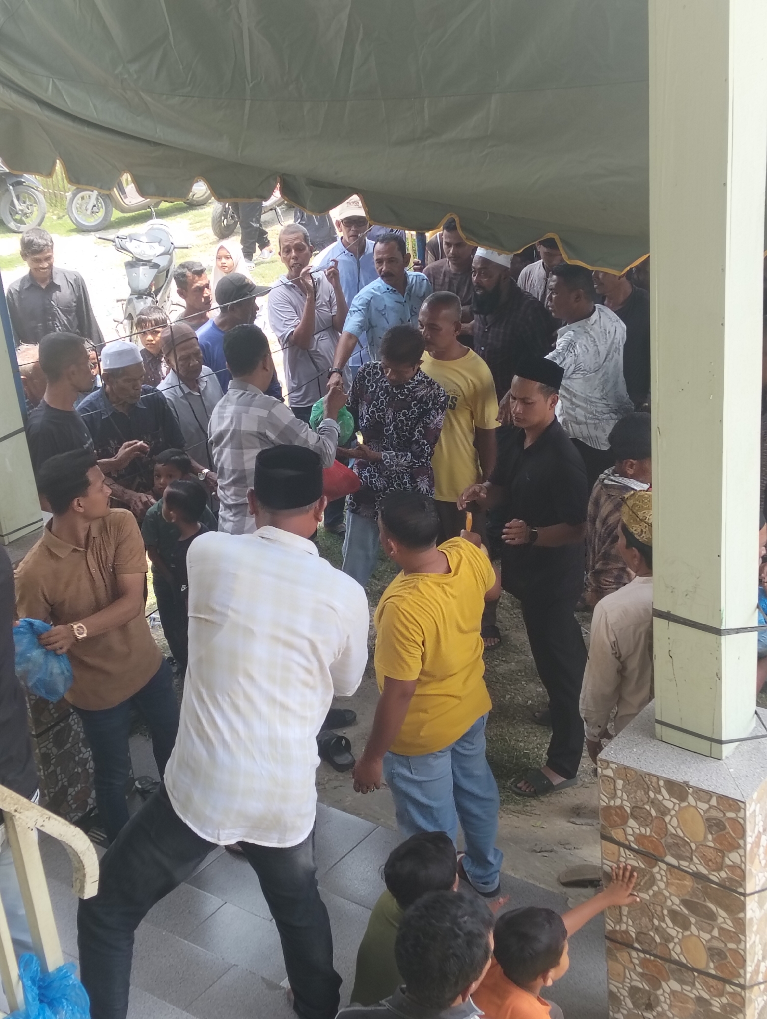 The image captures a gathering of people, mostly men, under a large tent in an outdoor setting. The scene appears to be in the city of Grong-Grong, Indonesia, suggested by the attire of the individuals, which includes peci caps and batik-like patterns on some clothing. Several motorcycles are visible in the background, indicating a communal event or gathering.
In the foreground, a man in a white long-sleeved shirt and dark pants is facing away from the camera, his arms slightly bent as if in motion. To his left, a man in a brown shirt is holding a blue plastic bag and looking towards the left. Another man in a yellow t-shirt is visible to the right of the man in white, also with his back to the camera, wearing blue jeans.
A group of men are clustered in the middle ground, some conversing and gesturing. One man in a black polo shirt and black pants stands out, looking down with his hands held together. Another man in a grey checkered shirt is to his right. Several children are also present, some standing near the adults.
The environment is somewhat rustic, with a gravelly ground visible in parts. The tent provides shade, suggesting a warm climate. The overall atmosphere seems to be one of a community event, possibly a religious gathering or a local celebration.