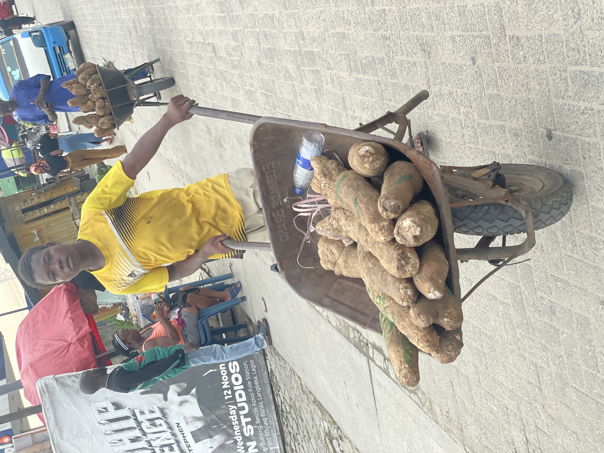 The image captures a vibrant street scene, likely a market, in Langbasa, Lagos, Nigeria, during daylight hours. The central figure is a young man, dressed in a yellow Adidas t-shirt and light trousers, who is pushing or standing with a rusty wheelbarrow heavily laden with numerous large, brown yams. Inside the wheelbarrow, a clear plastic CWAY water bottle and a pink measuring tape are visible. The man looks directly at the camera with a neutral expression.
In the background, another man in a blue outfit is seen next to a second wheelbarrow, also filled with yams, suggesting a trade in agricultural produce. A blue bus or truck is partially visible further back. Several other people are scattered throughout the scene; one individual is seated under a red umbrella on the left, and two others are near a large banner. The ground is paved with light-colored bricks.
The prominent banner on the left provides several location cues and details, including "Wednesday | 12 Noon," "N STUDIOS," "Say Building, Beside Azkol Fule Station, Adjacent Sterling Banks, Langbasa, Lagos." These details specify the time, a studio, and local landmarks, confirming the location as Langbasa, Lagos. The bright lighting suggests it is sunny. The overall activity depicts a bustling commercial environment where yams are being sold or transported.