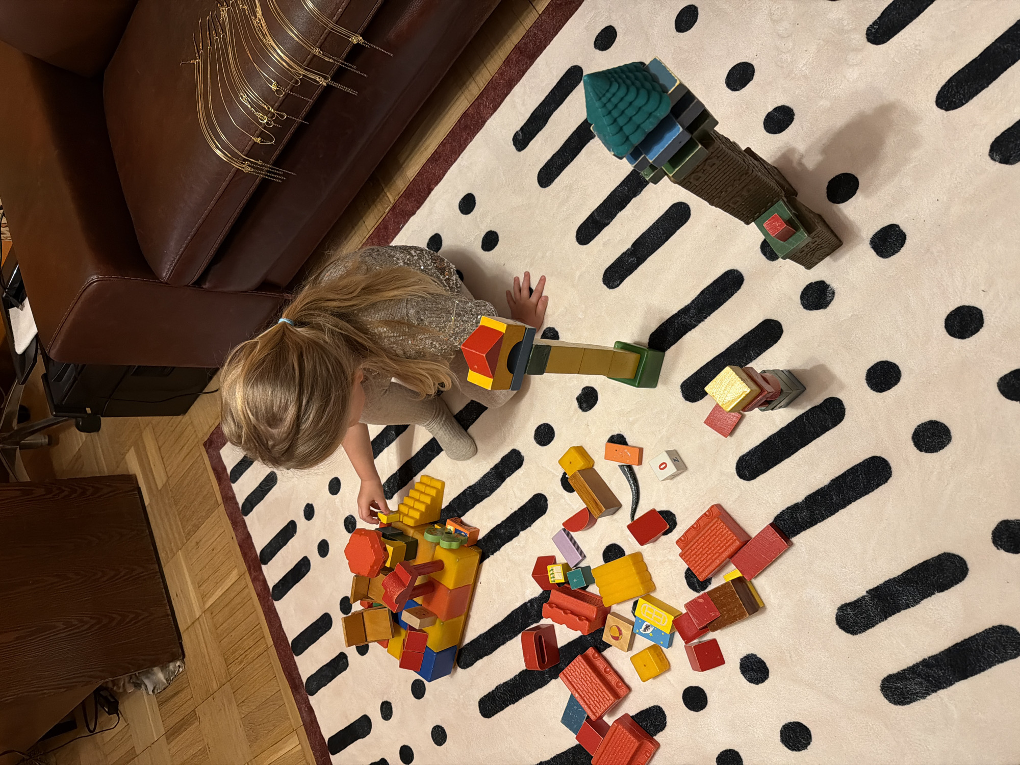 An aerial, slightly tilted view captures a young child engrossed in play on a patterned rug indoors. The child, likely a girl with blonde hair tied in a ponytail, is seen from the back, wearing a sparkly top and light-colored pants. She is kneeling on a light-colored rug featuring a distinctive graphic pattern of black dots and thick black dashed lines.
The primary activity involves building with a diverse collection of colorful wooden blocks. Several constructions are visible: a tall, intricate tower-like structure with a green pyramidal top stands in the upper right portion of the rug. Near the child's left hand, a more complex, multi-colored assembly with various pegs is taking shape. Numerous individual blocks in shades of red, yellow, blue, green, and brown are scattered across the rug, indicating an ongoing and active play session. A small white block has the number "0" and the letter "N" printed on it, while a yellow block features a small, indistinct blue and white graphic.
The setting is an indoor environment, likely a home in Khimki, Russia. A section of a brown leather sofa or armchair is visible in the upper left, draped with golden, chain-like decorative items. The floor surrounding the rug is made of dark wooden planks, possibly parquet or laminate. The lighting suggests an indoor setting, likely artificial. The child appears deeply absorbed in the creative process of building with the blocks.
