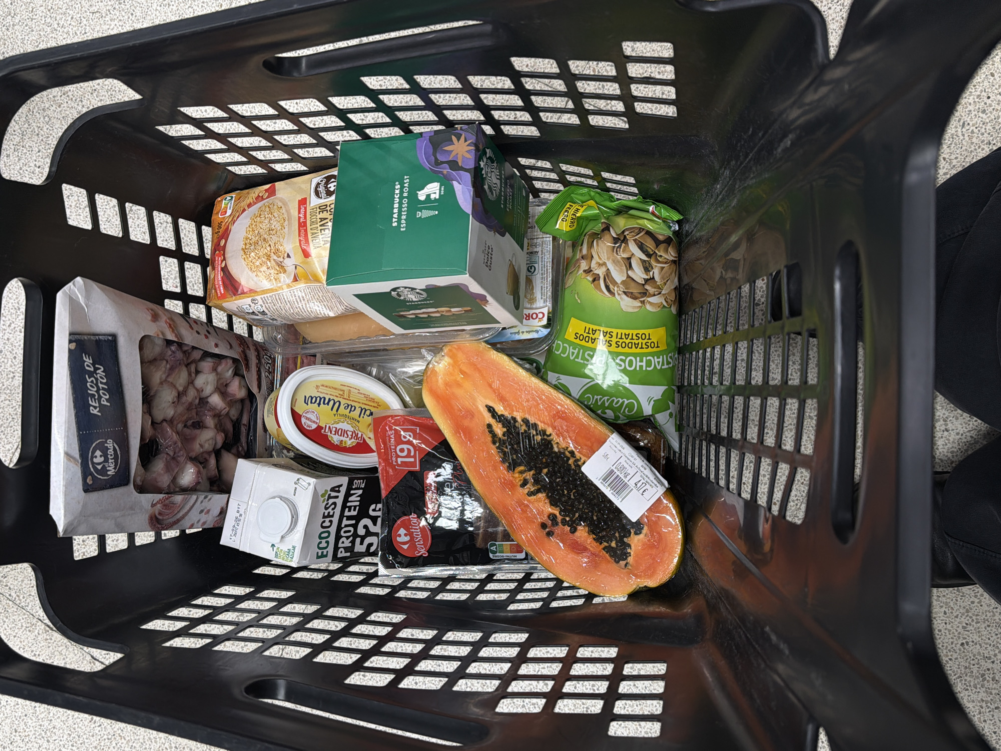 The image shows a black plastic shopping basket filled with various grocery items. The items are: a package of squid pieces ("Rejos de Potón"), a carton of Ecocesta protein drink, a container of President brand spreadable butter, a package of Sensation brand protein snack, half of a papaya wrapped in plastic, a box of Starbucks Espresso Roast Dolce Gusto capsules, a bag of roasted salted pistachios, and a package of oat flakes. The items are packed closely together in the basket. The basket has a grid-like pattern in its design. A person's leg is visible at the right side of the image. The setting appears to be a supermarket or grocery store, with a gray-beige floor pattern visible beneath the basket. The products indicate that the location is likely in Spain. There is no explicit evidence linking this image to the city of Santiago De Compostela.