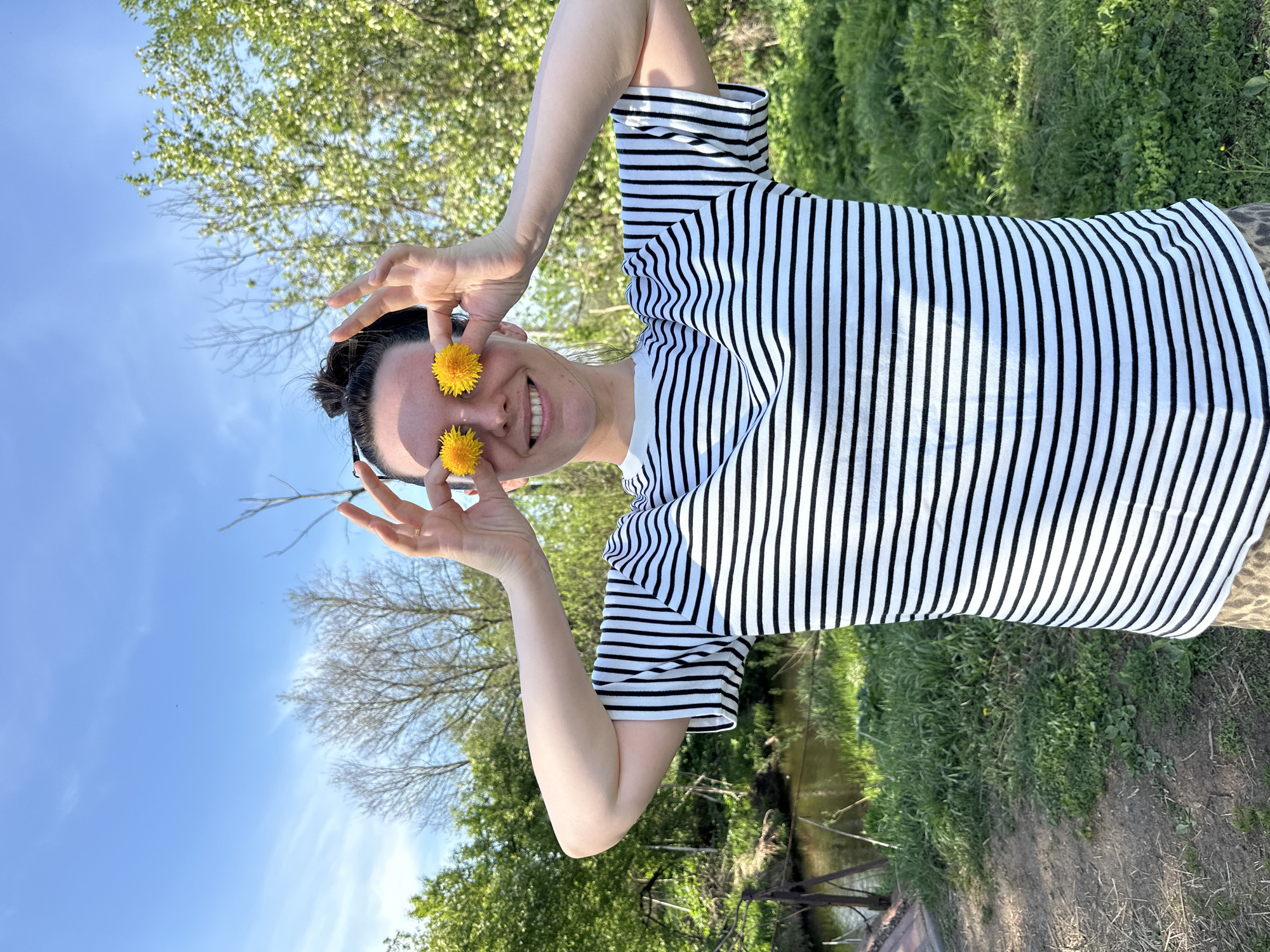 A person is holding two yellow dandelion flowers over their eyes while outside. They are wearing a white and black striped shirt. The background features a blue sky, trees, and greenery. The location appears to be a park or grassy area. The city of Альняш, Russia is not discernible from the provided details in this image.