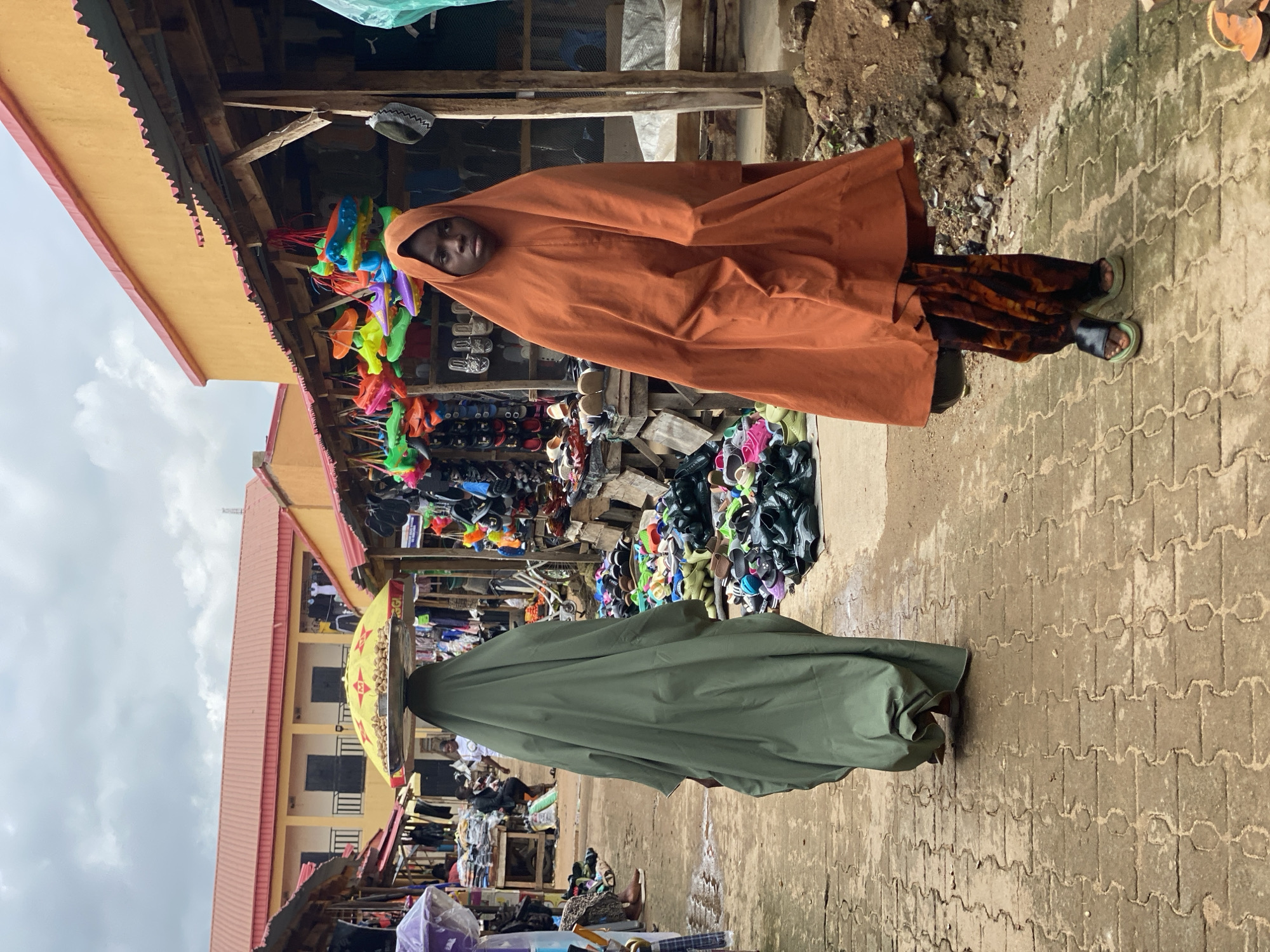 In Kafanchan B, Nigeria, a marketplace scene unfolds under a cloudy sky. Two women, both draped in traditional attire, are prominent. One, in the foreground, wears a striking orange hijab and a patterned skirt, her gaze directed forward. Behind her, a second woman is seen from the back, covered in a long, olive-green garment. She carries a large, yellow umbrella balanced on her head, atop a tray laden with small, light-brown objects.
The environment is a bustling market with stalls lining a cobbled pathway. To the right, a wooden stall displays an array of brightly colored children's shoes hanging from above. Shelves below are filled with more footwear, predominantly sandals and flip-flops in various shades. To the left, a similar stall displays clothing and other wares. The ground is a textured, interlocking brick pattern, showing signs of moisture. The overall impression is one of a vibrant, active market day.