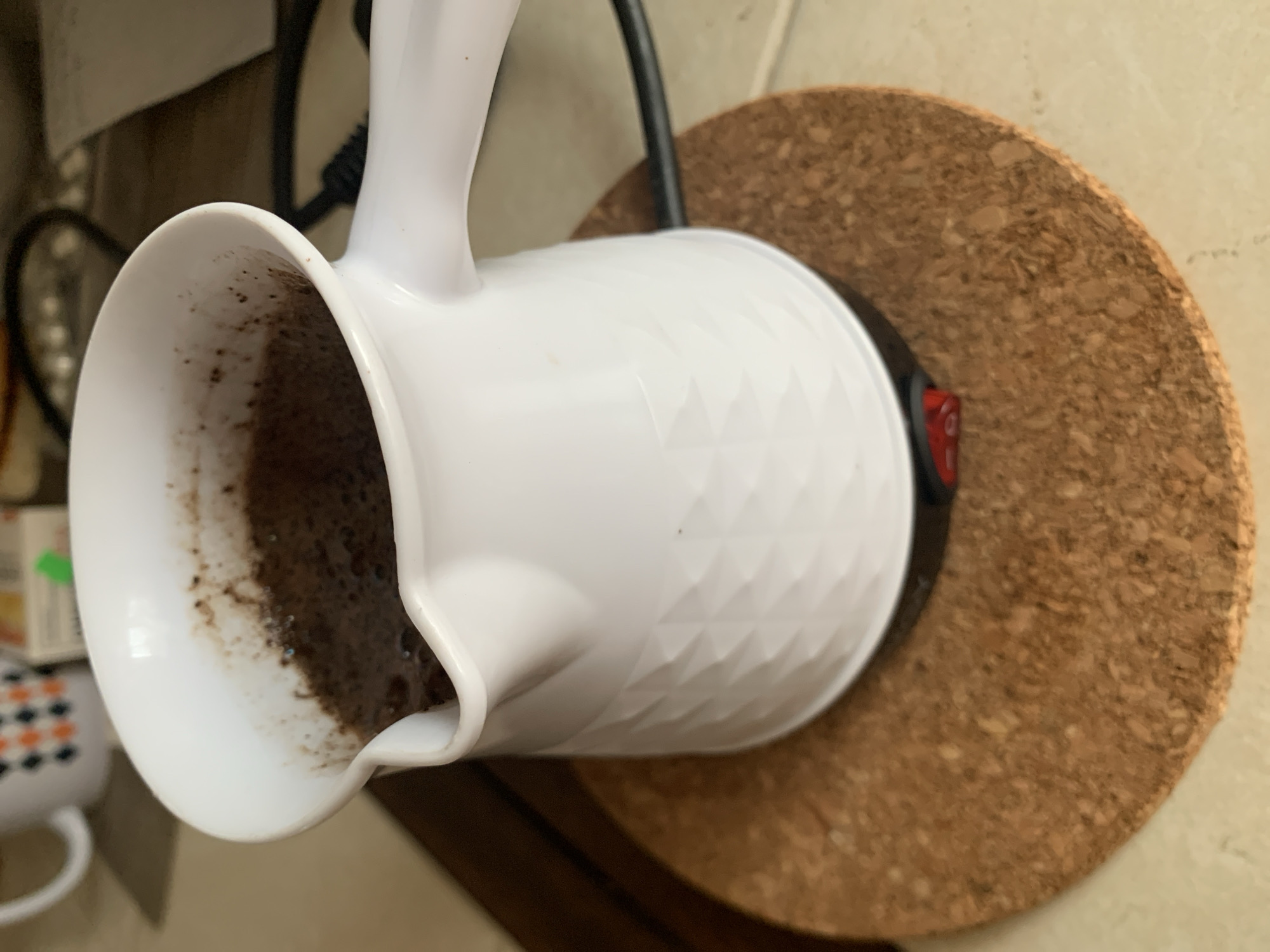 A white electric coffee maker with a diamond-patterned exterior sits on a cork coaster. The coffee maker is filled with dark coffee grounds and liquid. A red power switch is visible on the base of the coffee maker. The coffee maker is plugged into a black cord that disappears out of frame. The background shows a blurred countertop and a portion of a mug with an orange and black diamond pattern. The scene appears to be indoors, possibly in a kitchen. There are no people visible in the image. The location is not specified in the image.