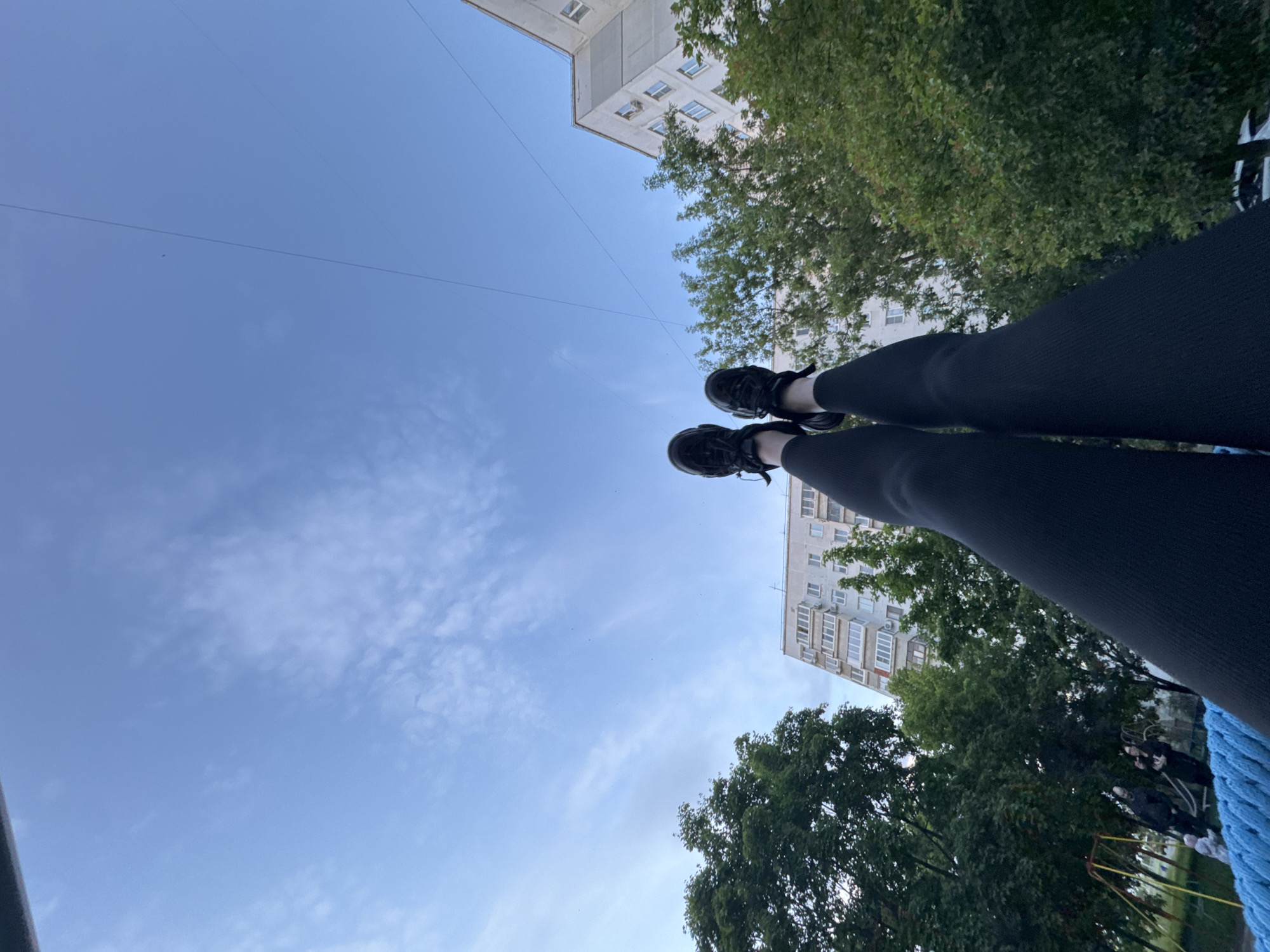 This image is taken from an extreme low-angle perspective, looking directly upwards towards the sky. In the foreground, a person's legs and feet are prominently featured, wearing black athletic shoes and black leggings or pants, extending into the frame from the bottom. A textured blue fabric or rope is visible in the bottom right corner, suggesting the photographer might be resting on a hammock or similar outdoor equipment.
The majority of the frame is dominated by a clear, bright blue sky with a few scattered, wispy white clouds. Faint, thin lines, likely utility wires, stretch across the sky. On both sides of the image, the upper sections of tall, light-colored apartment buildings with numerous windows and balconies are visible. These buildings are partially framed by abundant green trees with dense foliage, suggesting an urban park or residential area with significant greenery.
In the distant lower-right background, beyond the trees and partially obscured, several indistinct figures can be seen near what appears to be playground equipment, distinguished by yellow and red structural elements. The setting suggests a daytime scene, likely late morning or early afternoon, with fair weather. The overall impression is one of a relaxed moment spent outdoors in a residential urban environment. No text is visible in the image.