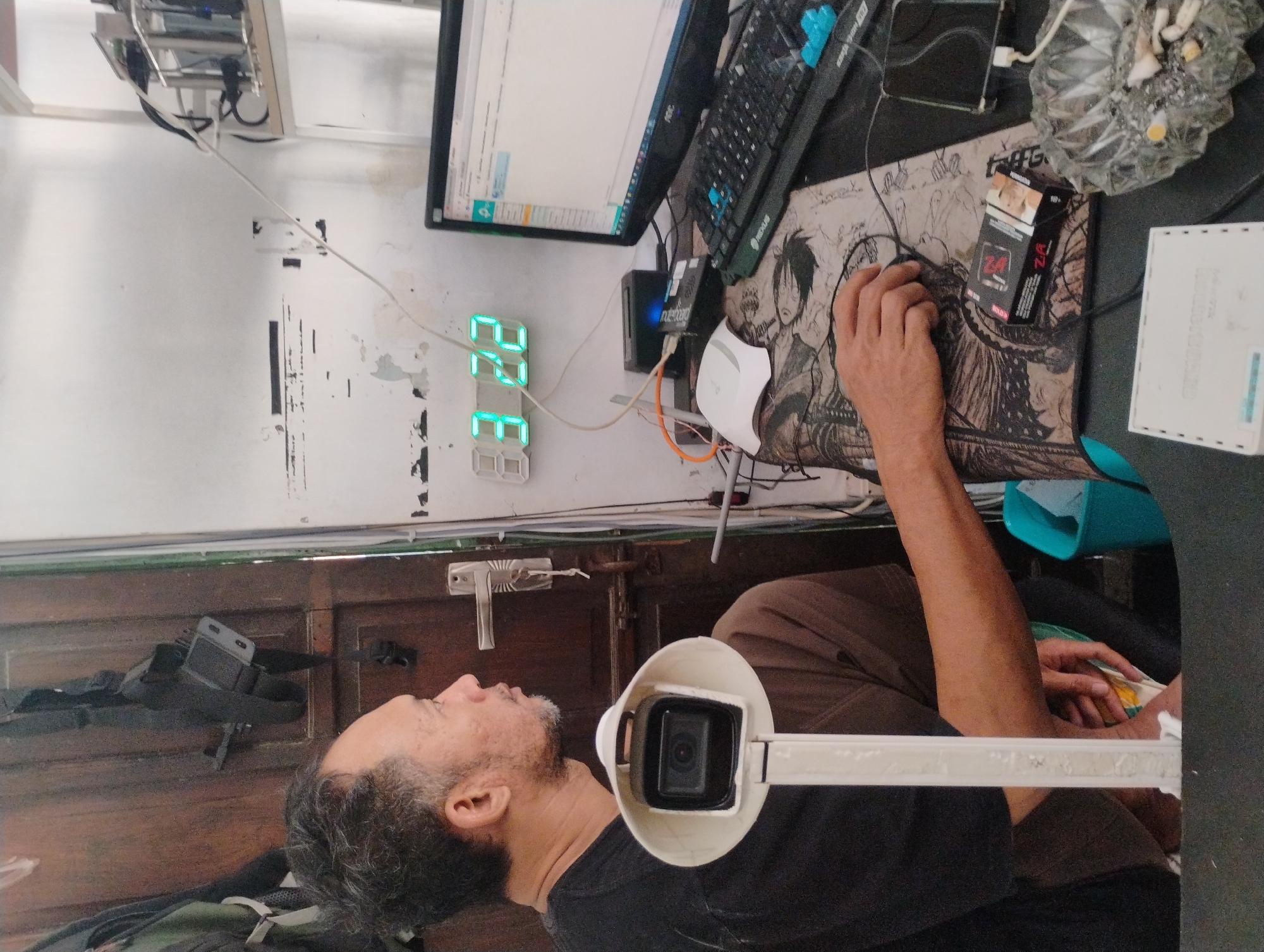 An Indonesian man sits at a desk with a computer and keyboard. A digital clock on the wall shows 03:22. A webcam is positioned in front of him. There is a pack of cigarettes and an ashtray with cigarette butts on the desk. The room appears to be cluttered with various electronic devices and cables. The scene is likely in a home or office in Nanggerang, Indonesia.