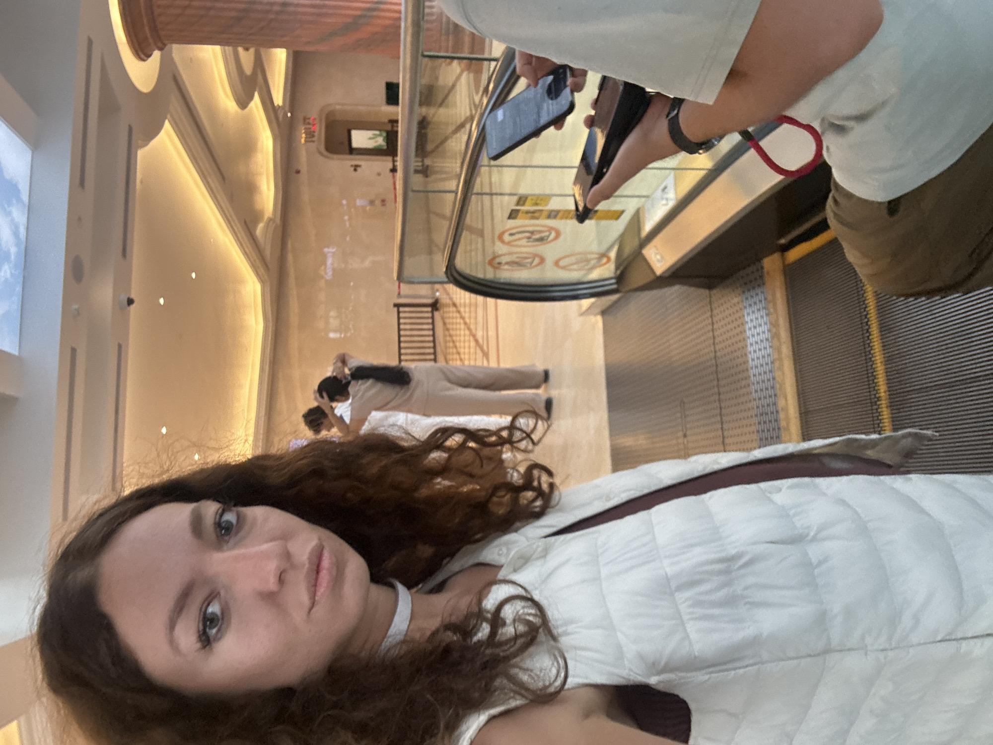 The image captures a selfie being taken by a young woman in an indoor, upscale setting, likely a mall or hotel in Đà Nẵng, Vietnam. The woman, positioned in the foreground and appearing to hold the camera for the selfie, has long, curly brown hair, blue eyes, and a neutral expression. She wears a white quilted vest over a darker garment and a white choker.
Behind her, to the right, another person is partially visible from the chest down, wearing a light-colored t-shirt and khaki pants. This person is also holding two smartphones over the glass barrier of an escalator, suggesting they might also be taking photos or videos.
Further in the background, several other individuals are present. Reflections on a wall show two people in light-colored attire, one appearing to pose or adjust their clothing, and another standing. These figures are seen near the base or on the lower section of the escalator.
The setting is characterized by grand architecture with warm, concealed lighting illuminating decorative curved and layered wall panels. A large, fluted column is partially visible at the top. An escalator with glass side panels and a black handrail occupies a significant portion of the right side of the frame. The escalator steps show metallic grating.
Notable details include:
* **Visible text:** The word "TOILET" is visible in red, vertical lettering on a sign in the background. On the escalator's glass barrier, several circular warning symbols (e.g., no running, no leaning) are present, along with some unreadable text.
* **Lighting:** The environment is brightly lit with artificial warm light, suggesting an indoor scene regardless of the actual time of day.
* **Activity:** The primary activity appears to be casual photography or selfies within a public building.