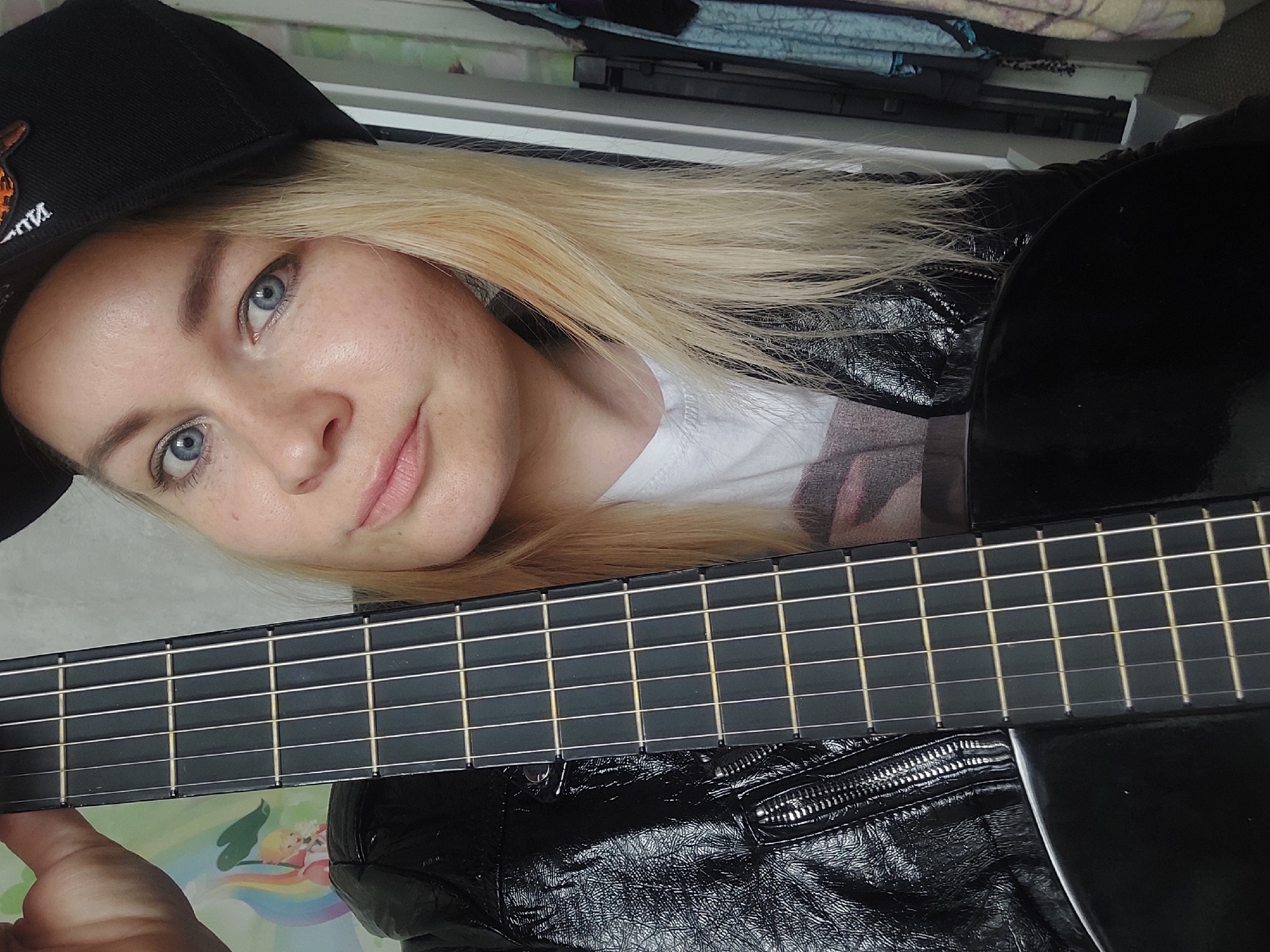 The image depicts a fair-skinned woman with blonde hair and blue eyes, wearing a black baseball cap and a shiny black jacket. She is holding a black guitar in front of her. The scene appears to be indoors, possibly in a home setting, with a glimpse of furniture and belongings in the background. There's no indication of the location being specifically in the city of Chaikovsky, Russia. The image seems to be a selfie taken by the woman, focused on her face and the guitar.