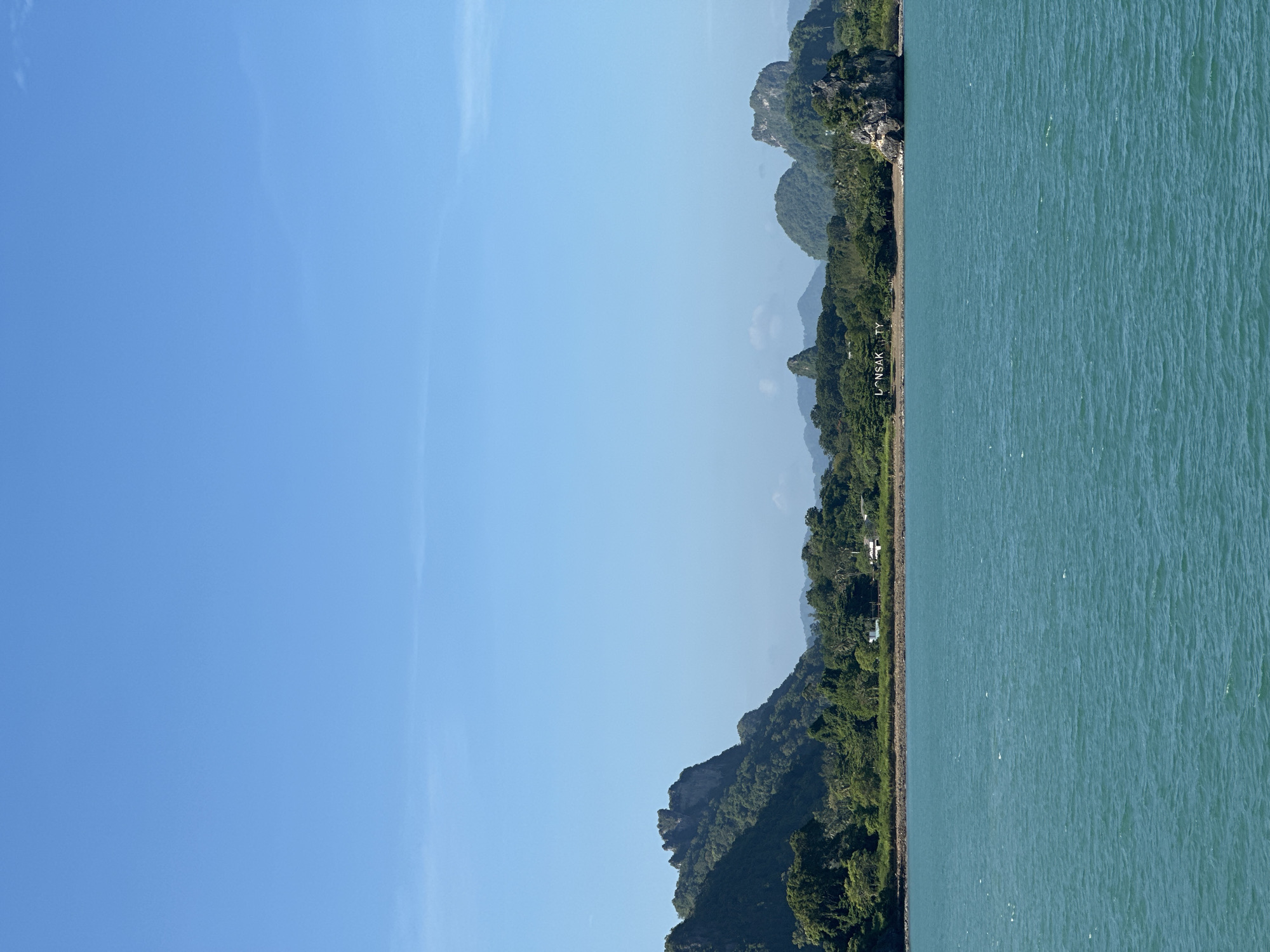 The image shows a wide view of the Gulf of Thailand, with the coastline of Don Sak, Thailand, in the background. The water is a vibrant turquoise, with small ripples indicating a gentle breeze. The shoreline is lined with lush green trees and vegetation, giving way to rolling hills and dramatic, rocky mountains. Several buildings, including what appears to be a white structure with a red roof, are visible nestled amongst the trees. In the mid-ground, large white lettering spelling out "LONSAK TY" is visible on the coastline, likely a landmark or sign. The sky is a clear, bright blue with a few wispy clouds near the horizon. The overall scene depicts a tropical coastal landscape.