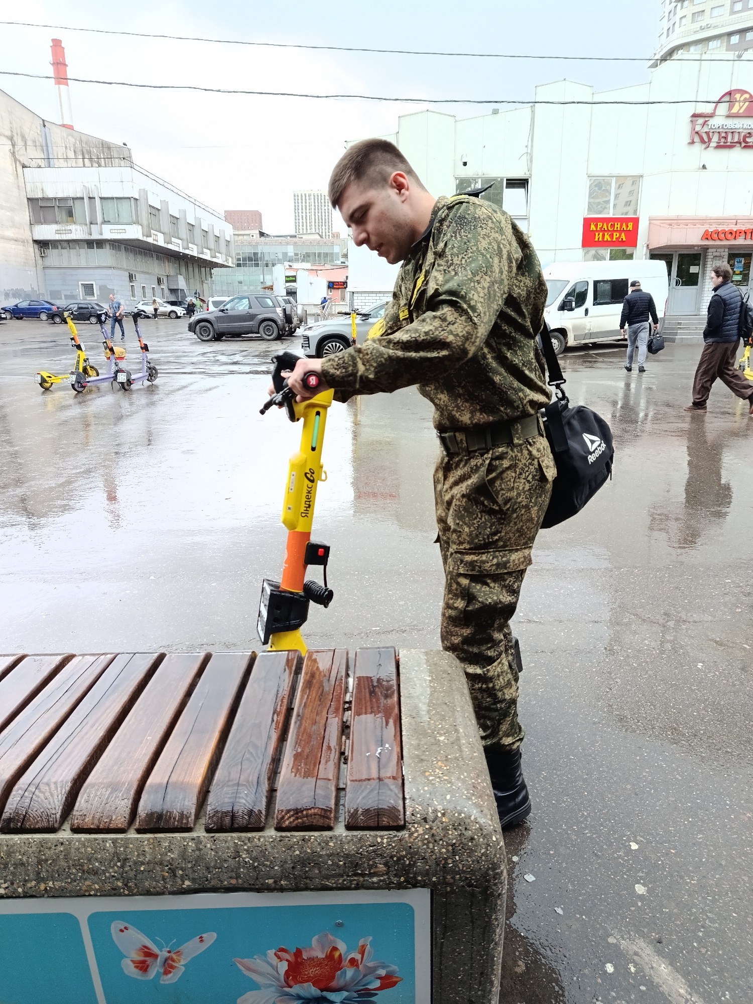 A man dressed in military fatigues stands on a wet city sidewalk in Moscow, Russia, with a yellow Yandex Go electric scooter next to a wet bench. The man is looking at the scooter's handlebars. He is wearing a black Reebok bag slung over his shoulder. In the background, there are several parked cars, a white van, and a building with the words "КРАСНАЯ ИКРА" (red caviar) and "АССОРТИ" (assortment) in red letters above an entrance. Other pedestrians are visible in the background, and a red-and-white smokestack is partially visible above a building. Several other rental electric scooters are parked nearby. The pavement is wet, reflecting the overcast sky.