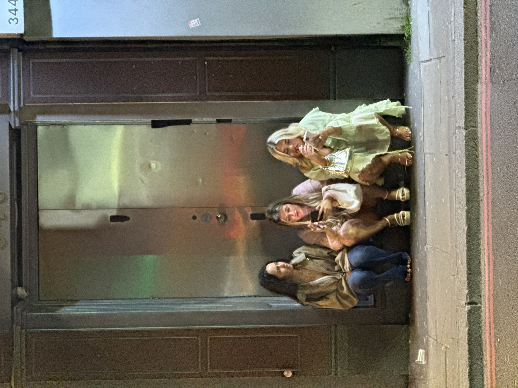 Three women are sitting on the sidewalk in front of a building at 344 Old Street, London, United Kingdom. The building has a metallic door and brown accents. The sidewalk is grey, with a red and white painted curb. The first woman is blonde and wearing a light green dress, gold shoes, and holding a silver clutch. She is looking at her phone. The second woman has dark hair and is wearing a light pink shirt and tan shorts. She is also holding a phone. The third woman is wearing a tan jacket and blue jeans. There is a clear plastic cup on the sidewalk near her.
