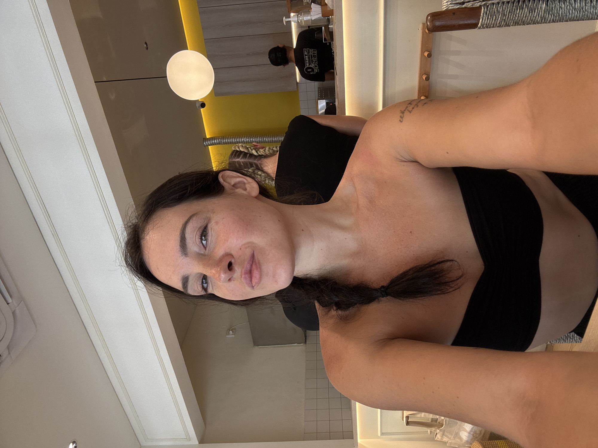 This is a portrait of a woman in North Kuta, Indonesia, taken from a selfie perspective. She is indoors, with a bright yellow wall visible in the background, suggesting a modern and perhaps casual setting like a cafe or restaurant. A circular white light fixture hangs from the ceiling. In the background, a person wearing a black t-shirt with text is visible behind a counter, likely working. The woman has dark hair styled in a braid and is wearing a black top. Her expression is relaxed and she has a small tattoo on her arm. The image appears to be taken during the daytime.