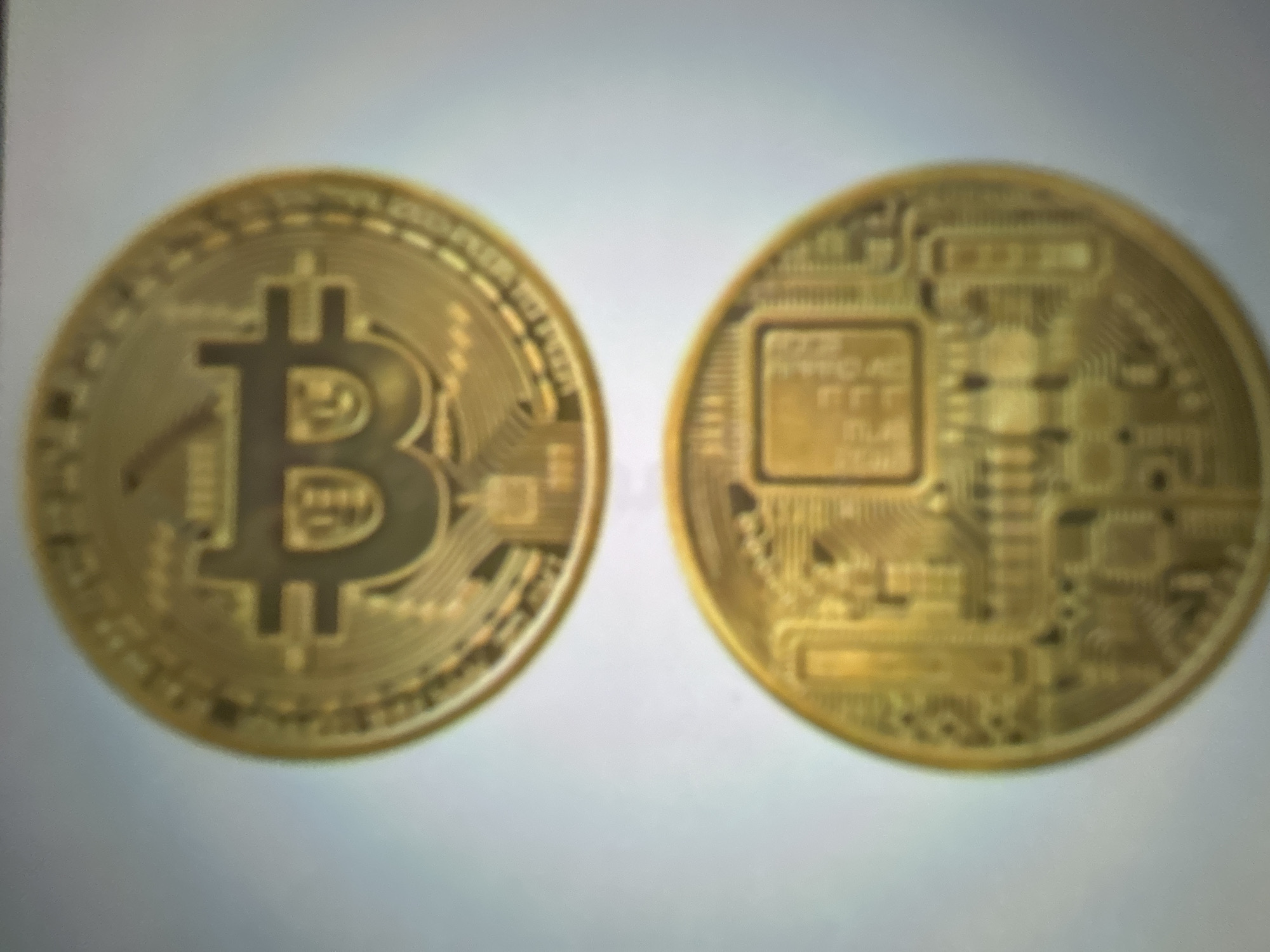The image displays two golden Bitcoin coins placed side-by-side against a plain, light-colored background. The left coin prominently features the Bitcoin logo, a stylized "B" with vertical lines, encircled by decorative patterns and text. The right coin exhibits a more intricate design, resembling a circuit board with various squares and lines, also with surrounding decorative elements and text. The image is somewhat blurred, making it difficult to discern fine details or read any inscriptions clearly. The lighting appears to be neutral, without strong shadows, suggesting an indoor setting or diffused light. There are no people, specific location cues, or indications of time or weather. The overall impression is a close-up view of collectible or novelty Bitcoin coins, emphasizing their metallic sheen and symbolic imagery.