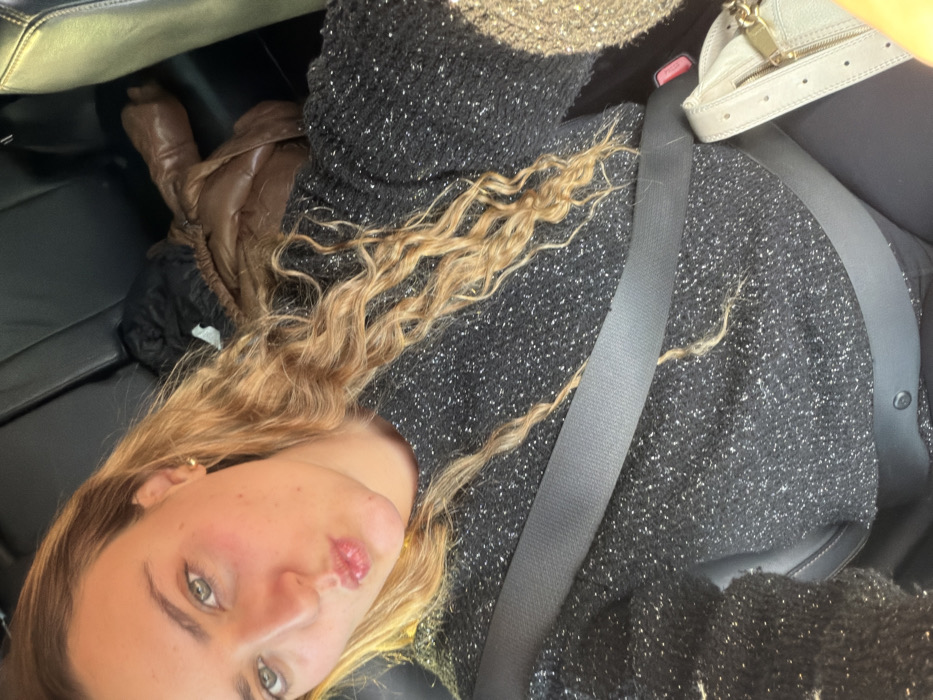 A selfie taken inside a car features a woman with curly blonde hair, green eyes, and pink lips. She's wearing a sparkly black top and a seatbelt. In the background, part of a brown and black jacket is visible on the leather car seat. A beige purse is also visible, partially cut off by the frame. The angle is tilted, creating a casual and intimate feel.