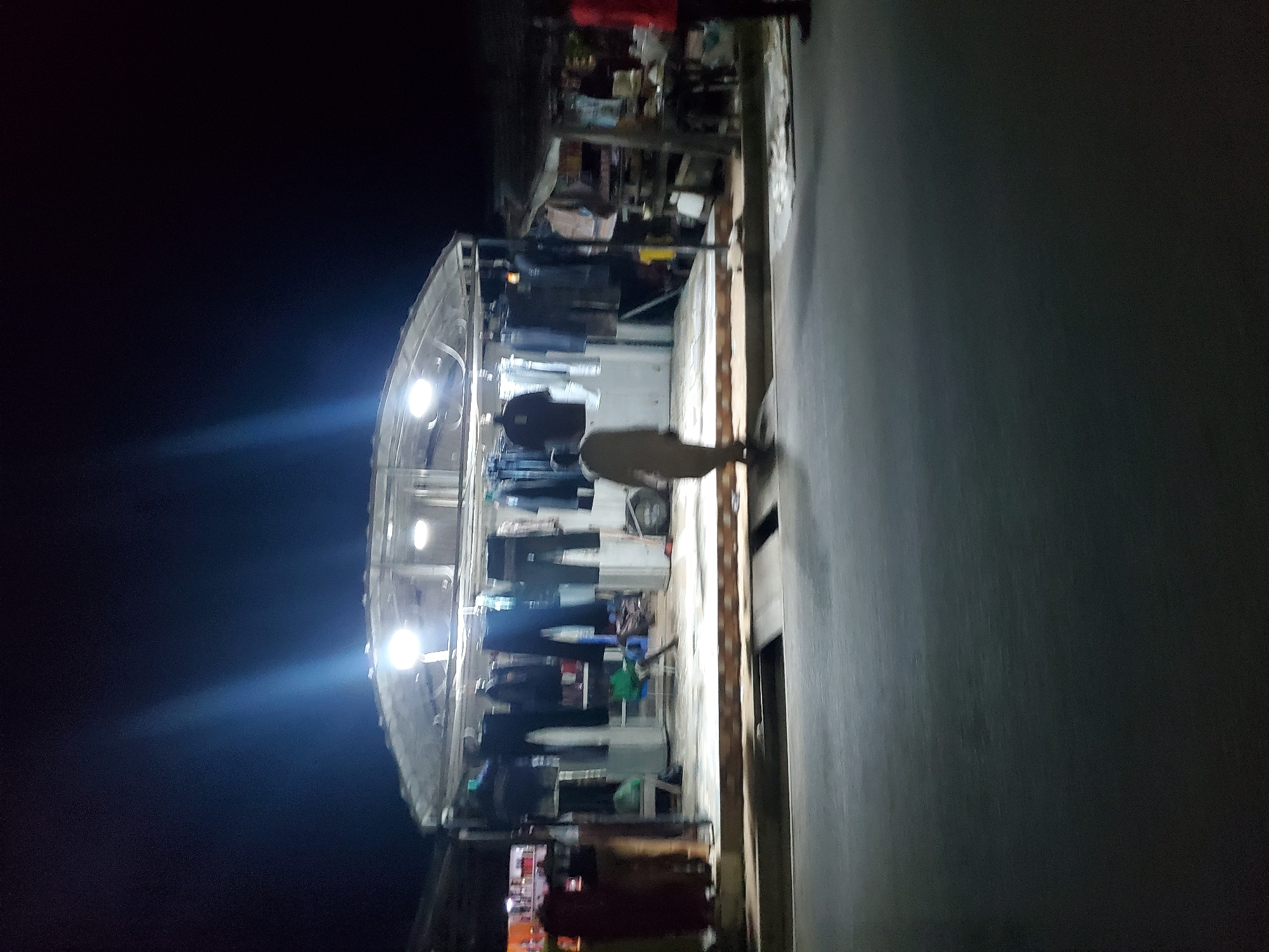 The image depicts an outdoor market scene at night in Jalingo, Nigeria. The primary subject is a market stall illuminated by overhead lights, displaying a variety of clothing items hanging on racks. The stall has a covered roof, suggesting protection from the elements. In the foreground, a person is partially visible, facing away from the camera and appearing to walk along the path next to the stall. The path itself is paved and appears to be a street or walkway.
The scene is characterized by the darkness of nighttime, with the artificial lights of the stall creating bright spots and some glare. The overall mood is somewhat dim and perhaps bustling, given it's a market. There are no visible signs of specific weather conditions, but the nighttime setting is prominent. The presence of clothing for sale indicates a commercial activity.
Notable details include the way the clothes are displayed, suggesting a retail environment. The lighting creates a dynamic contrast between the brightly lit stall and the surrounding darkness. The person in the foreground adds a sense of human presence and activity to the scene. The location context of Jalingo, Nigeria, places this scene within a specific geographical and cultural setting. There is no visible text in the image.