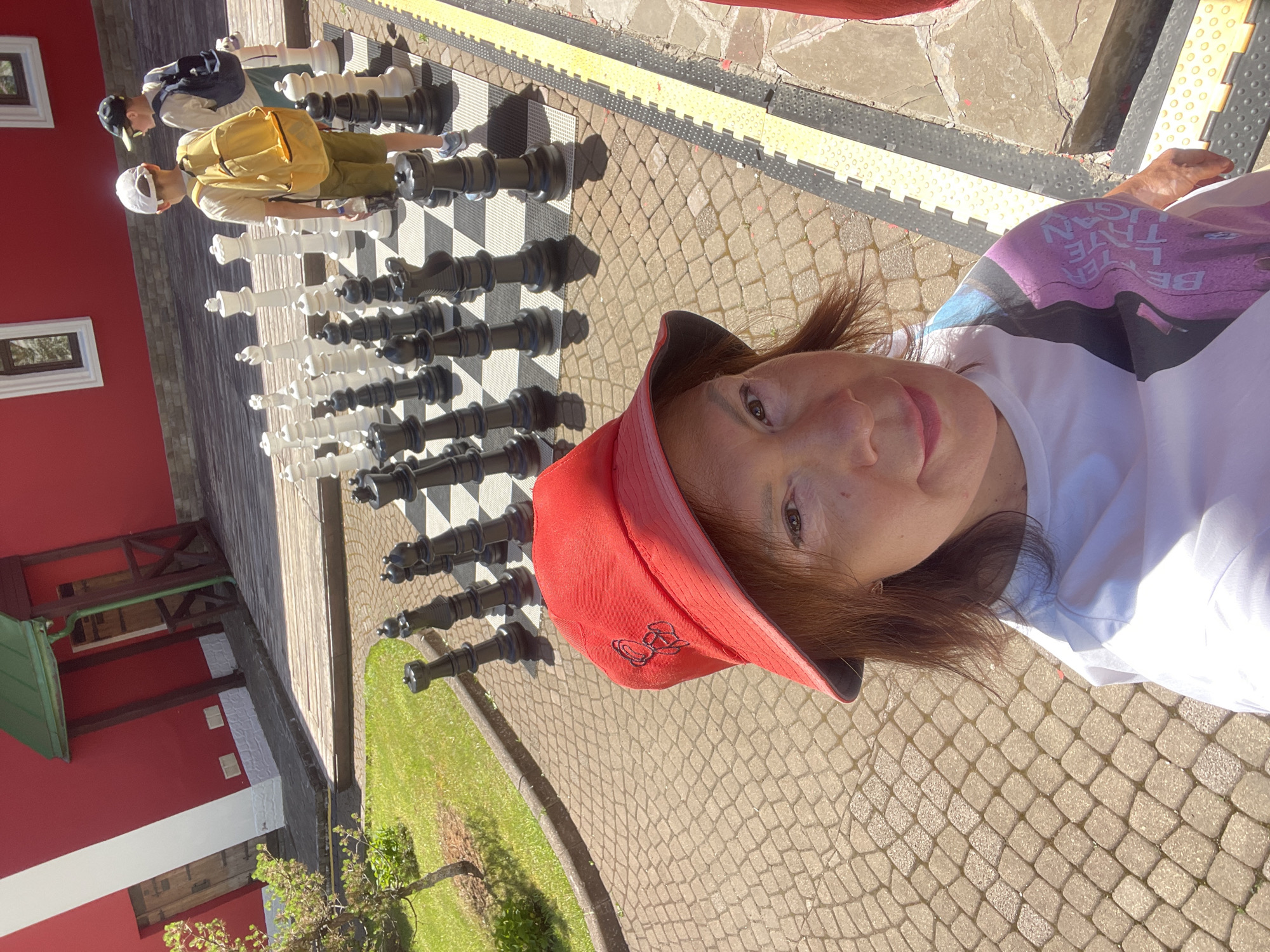In the city of Estosadok, Russia, a woman wearing a red bucket hat and a white t-shirt with a colorful graphic on the sleeve takes a selfie. She is standing on a paved area with cobblestone patterns, facing a giant chessboard set up outdoors. In the background, two young boys, one wearing a yellow backpack and the other a blue backpack, are positioned near the chessboard, appearing to be playing or observing. A bright red building with white trim and wooden elements is visible behind the chessboard. The scene is bathed in sunlight, casting shadows from the chessboard pieces onto the ground.