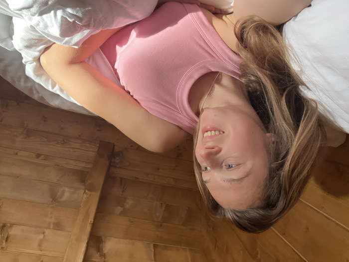In an interior setting within the city of Crkvine, Montenegro, a female individual is depicted lying in a bed. She is positioned on her back or side, with her head resting on a white pillow. Her long, light brown hair is loose around her head and shoulders. She wears a pink ribbed tank top and a thin silver-colored necklace. Her face is turned towards the viewer's right, with open eyes looking in that direction and a slight upward curve of the lips. Two faint, small, light-colored marks are visible above her left eyebrow. Her right arm is partially visible, resting on white bedding that covers the lower part of her body. The background consists of an inclined wooden ceiling or wall constructed from horizontal planks, with a prominent diagonal wooden beam. Strong natural light, consistent with direct sunlight, illuminates the scene from the right, casting distinct shadows.
