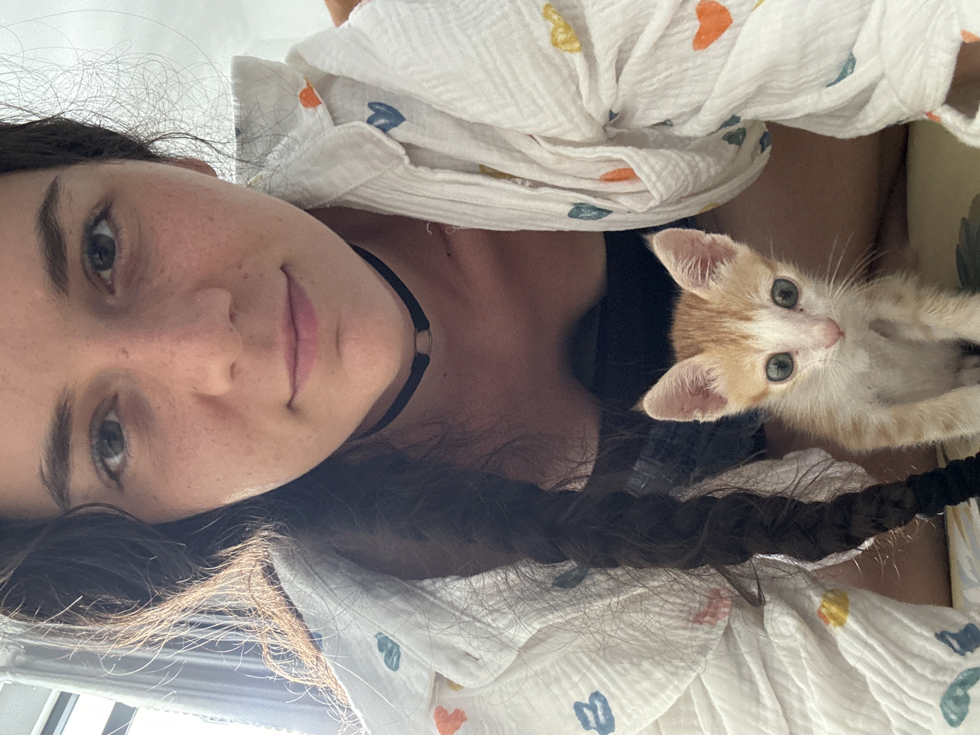 A person is lying down, cradling a light orange and white kitten. The person has brown hair with a braid that is visible. They are wearing a black choker necklace and a light-colored shirt with small colorful heart and flower designs. The kitten has light green eyes and appears to be looking directly at the camera. The setting is indoors, near a window providing natural light. The exact location in Mũi Né, Vietnam cannot be determined from the image alone.