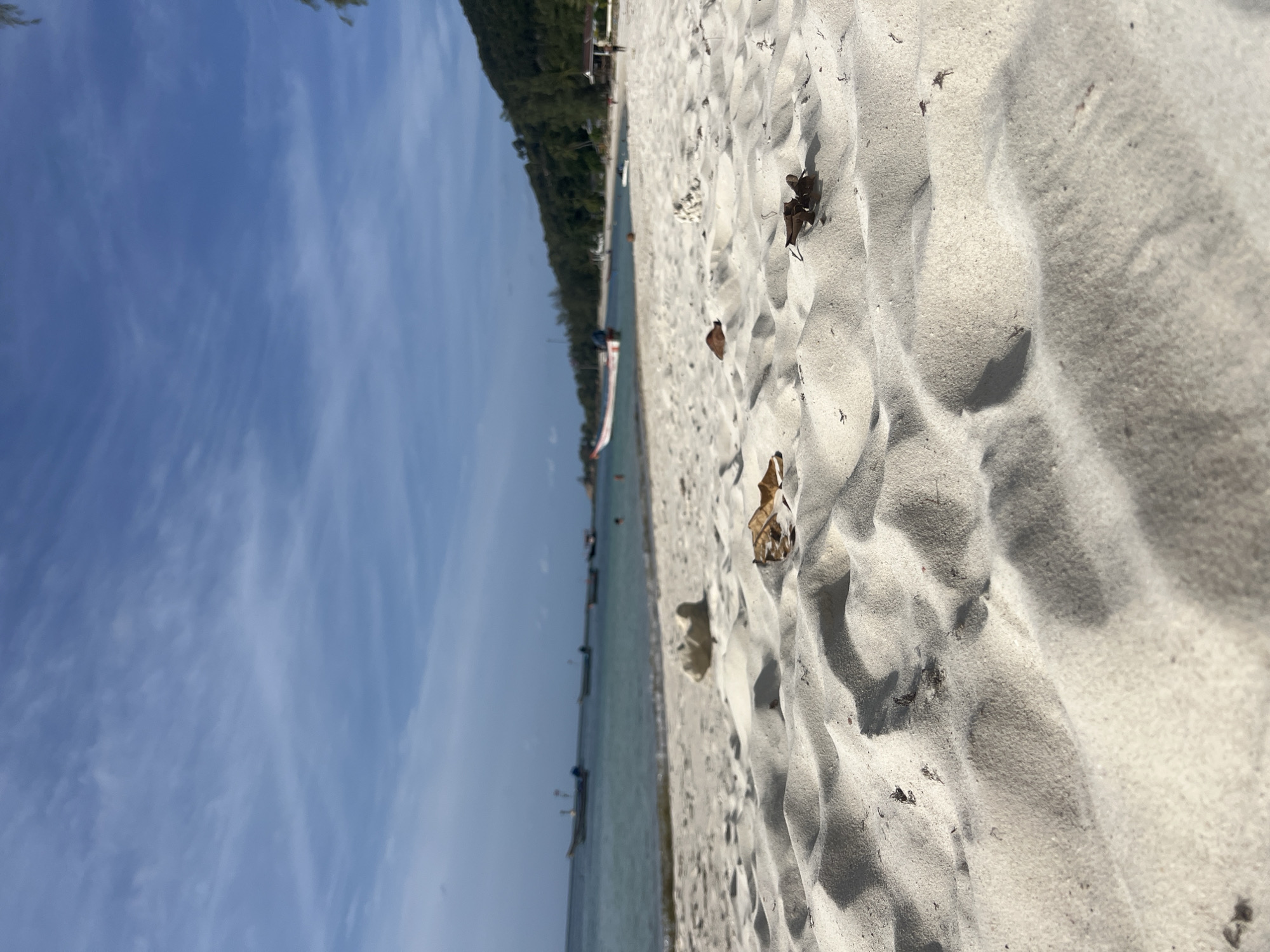 The image depicts a low-angle view of a sandy beach with the ocean in the background. The foreground is dominated by the texture of the white sand, with ripples and shadows creating a sense of depth. A few dry leaves are scattered on the sand. In the mid-ground, the shallow, calm turquoise water meets the shore. Several small boats are visible in the water, some anchored and others appearing to be moving. The horizon shows a line of green, tree-covered hills or land. The sky above is a clear blue with scattered white clouds. The scene suggests a tropical or coastal environment on a sunny day.