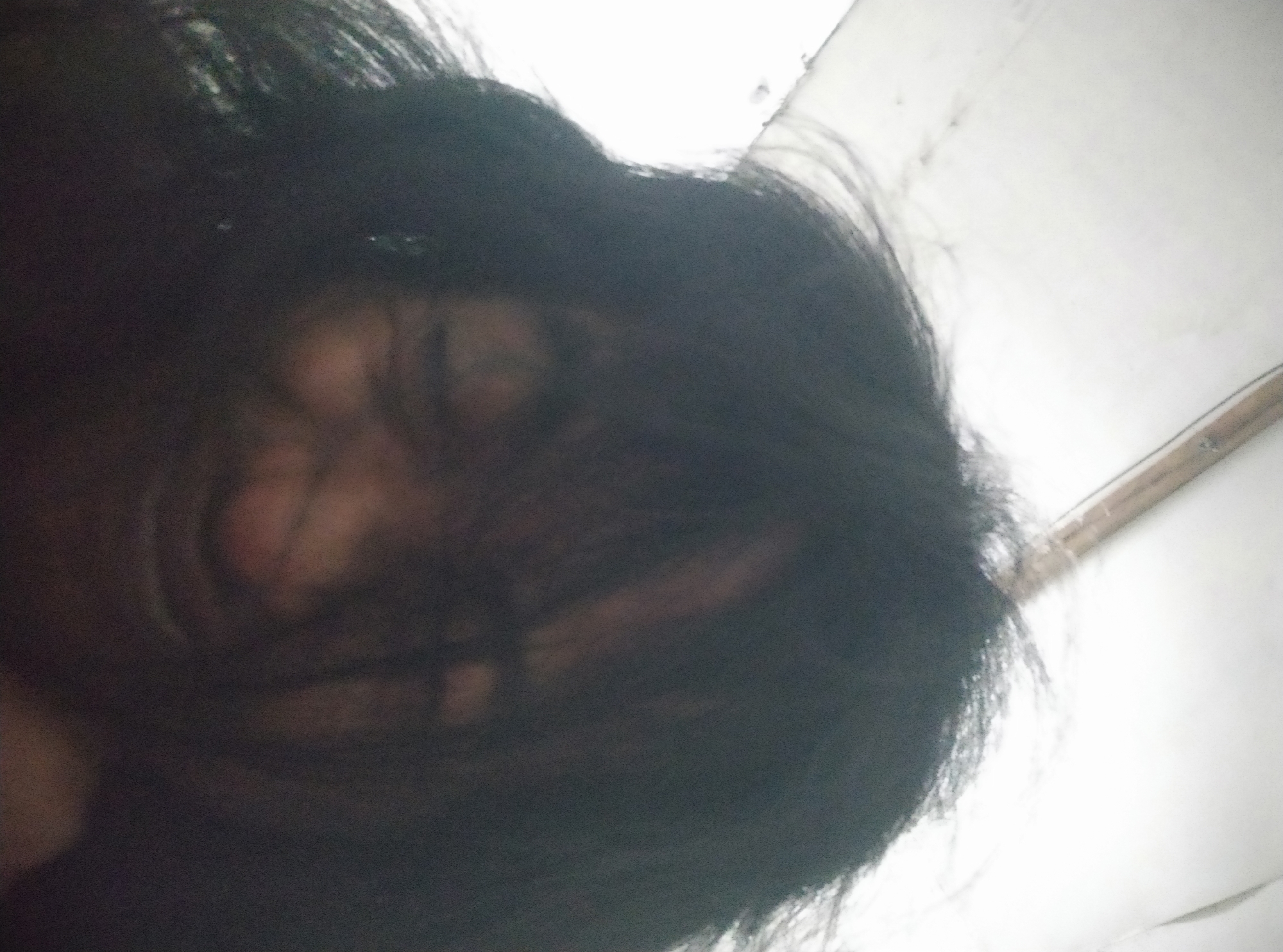 The image is a low-angle, close-up, and blurry selfie of a person with dark, messy hair. The person's face is partially obscured by their hair, with only hints of features visible. Their eyes appear closed or squinting, and their mouth is open in what could be a laugh or a grimace. The background is a bright, out-of-focus white surface, possibly a ceiling or wall. There is a thin, dark horizontal line across the top of the image. The lighting is harsh and uneven, creating strong shadows and highlights. The overall impression is one of informality and perhaps a candid moment. No specific location in Sumurber, Indonesia, can be identified from the image.
