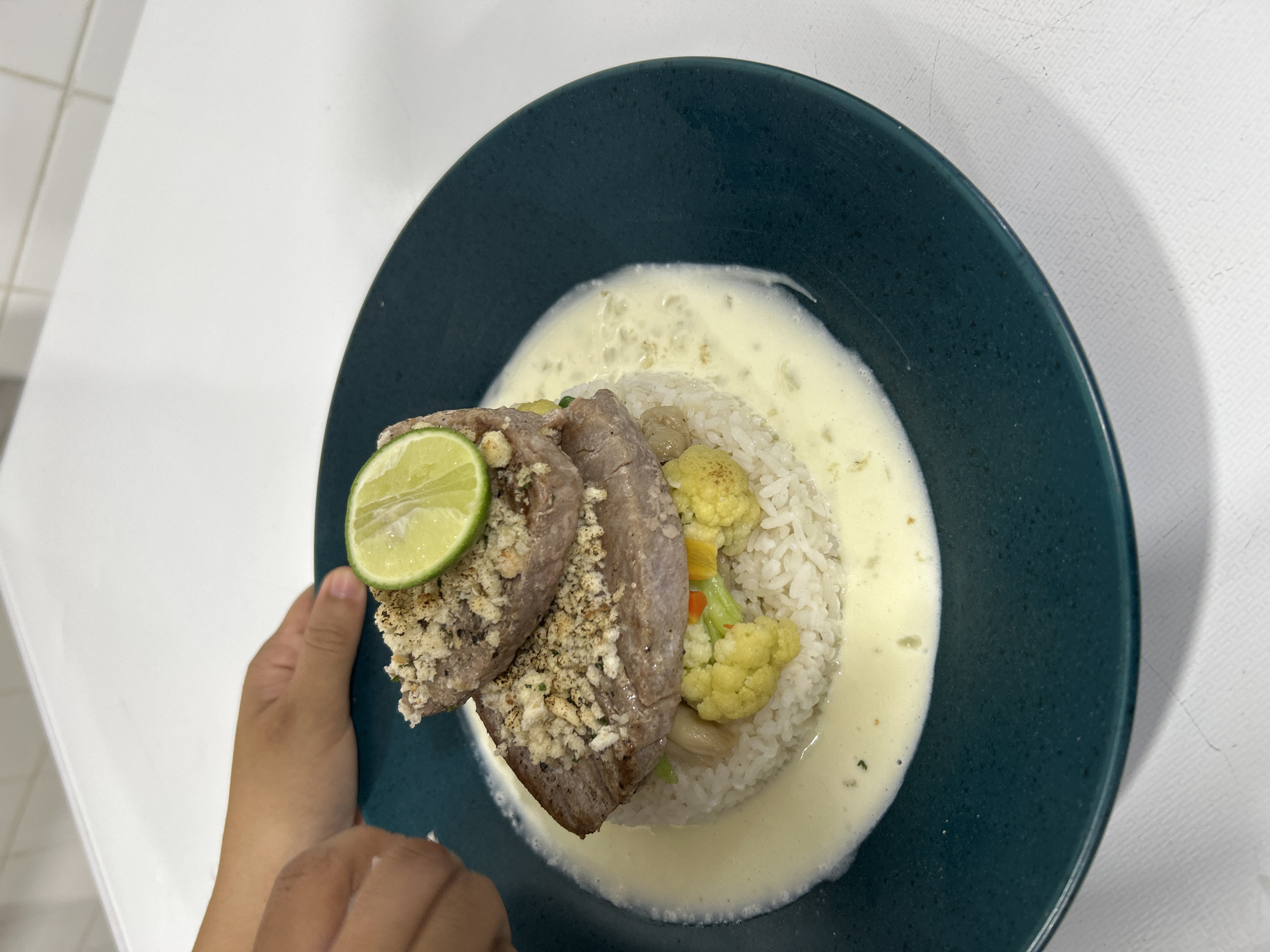 The image shows a plate of food held in a hand. The food appears to be a dish with rice, vegetables (including cauliflower), a creamy sauce, and what seems to be fish or meat, topped with a lime slice. The plate is dark green, and the background includes a white tiled wall and a white countertop or table. Given the location of Senggigi, Indonesia, it's likely that the dish includes regional ingredients or preparations common in Indonesian cuisine.