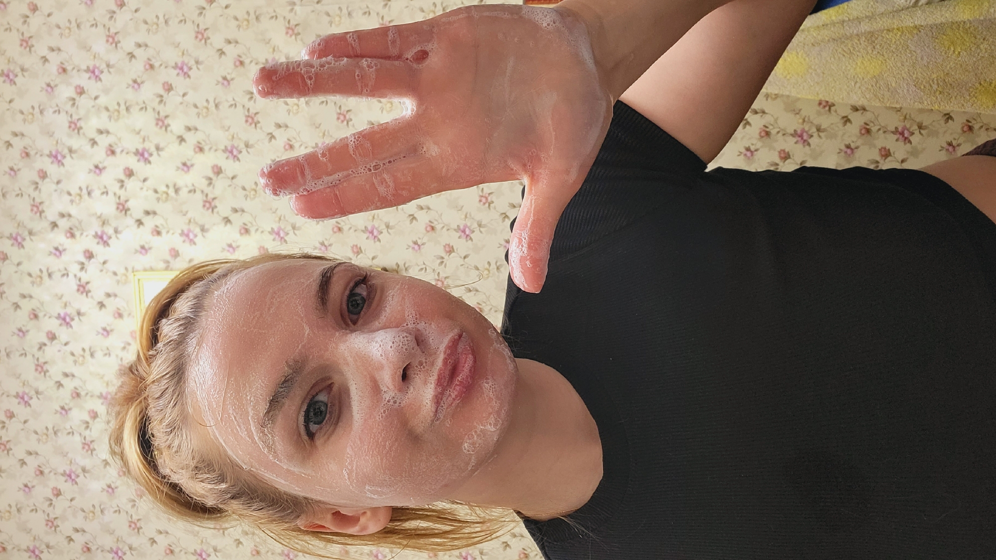 A woman with fair skin, blonde hair, and blue eyes is posing for a selfie. Her face and hand are covered in soapy bubbles. She is wearing a black shirt. The background is a light-colored floral wallpaper. The image was taken in Зипуново, Russia.