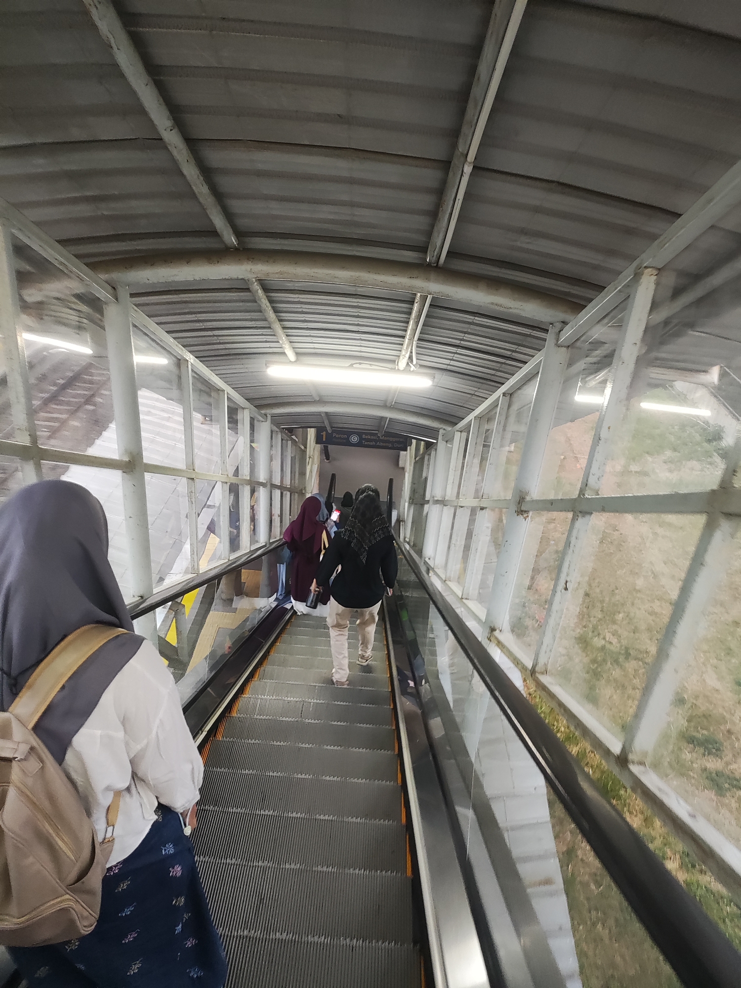 The image shows an outdoor escalator at a transportation hub, likely a train station, in Telagamurni, Indonesia. Several individuals are ascending the escalator, all wearing modest clothing, including headscarves. The escalator has a metal structure with glass walls, revealing dry, grassy terrain outside. The overall lighting suggests an overcast day or indoor setting. A sign with Indonesian text, "Peron 1 Stasiun Cikarang" and "Bekasi-Manggarai-Tanah Abang-Duri," indicates the destination and line. One person in the foreground is seen from behind, wearing a gray headscarf and a backpack, while others further up the escalator are partially visible. The scene is utilitarian and functional, typical of public transportation infrastructure.