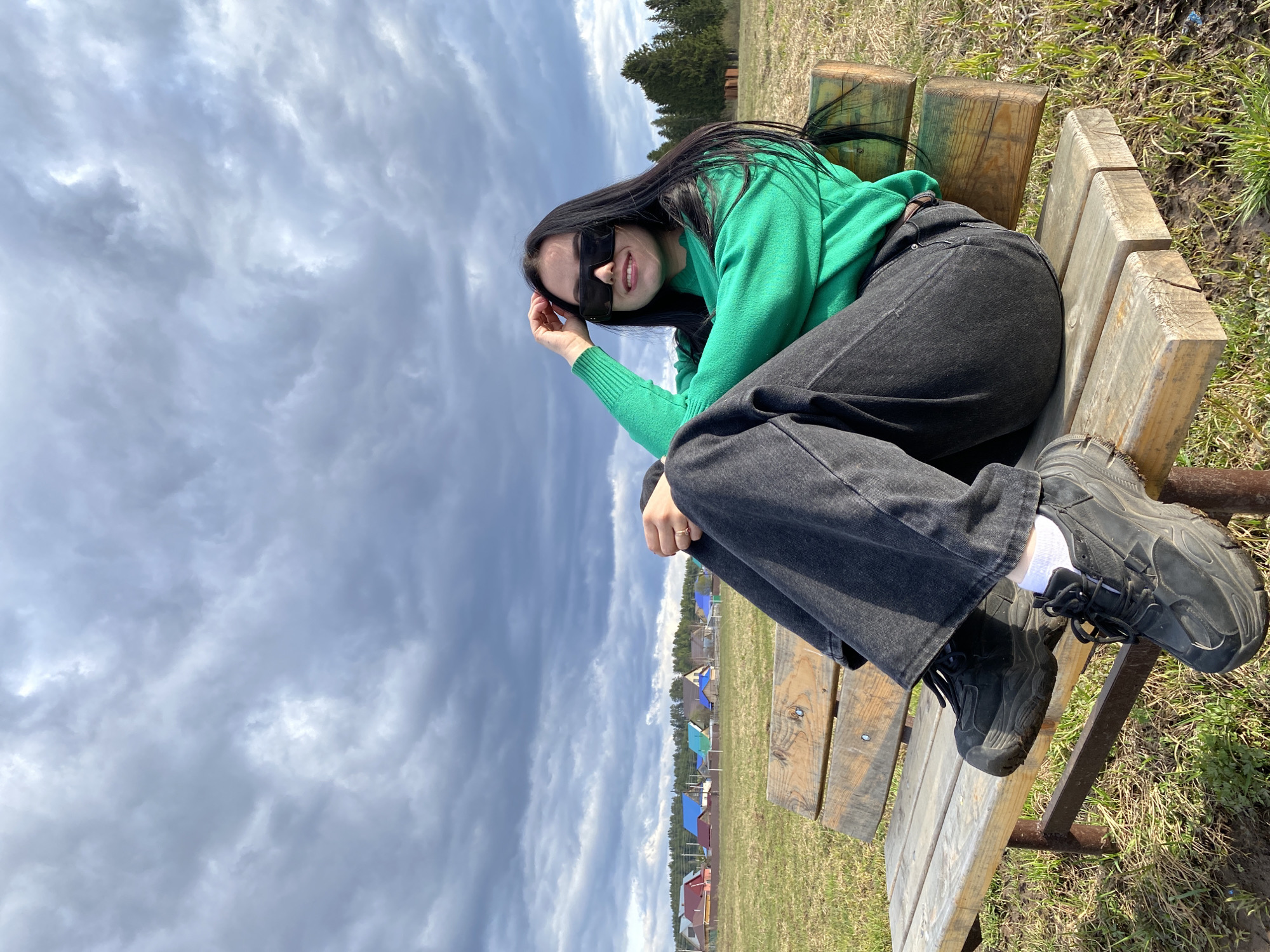 A young woman with long dark hair is captured in a dynamic, tilted shot, sitting on a weathered wooden bench outdoors. She wears a vibrant green long-sleeved sweater, dark wide-leg jeans, and chunky black sneakers with white socks. Dark rectangular sunglasses cover her eyes, but a subtle smile is visible on her face as she looks towards the camera, her right hand casually resting near her temple.
The setting appears to be a rural or semi-rural landscape, possibly in Foky, Russia, as suggested by the distant village. The background is dominated by a dramatic, heavily overcast sky with swirling grey and white clouds, indicating a cloudy or potentially stormy day. In the mid-ground, a line of dark green coniferous trees stands against the horizon. Below, a cluster of houses with distinctly colorful roofs, including blue, red, and green, forms a small settlement. The foreground features sparse, light green and brown grass, typical of early spring or late autumn. The woman's pose and expression convey a sense of relaxed contentment in the open air. There is no visible text in the image.