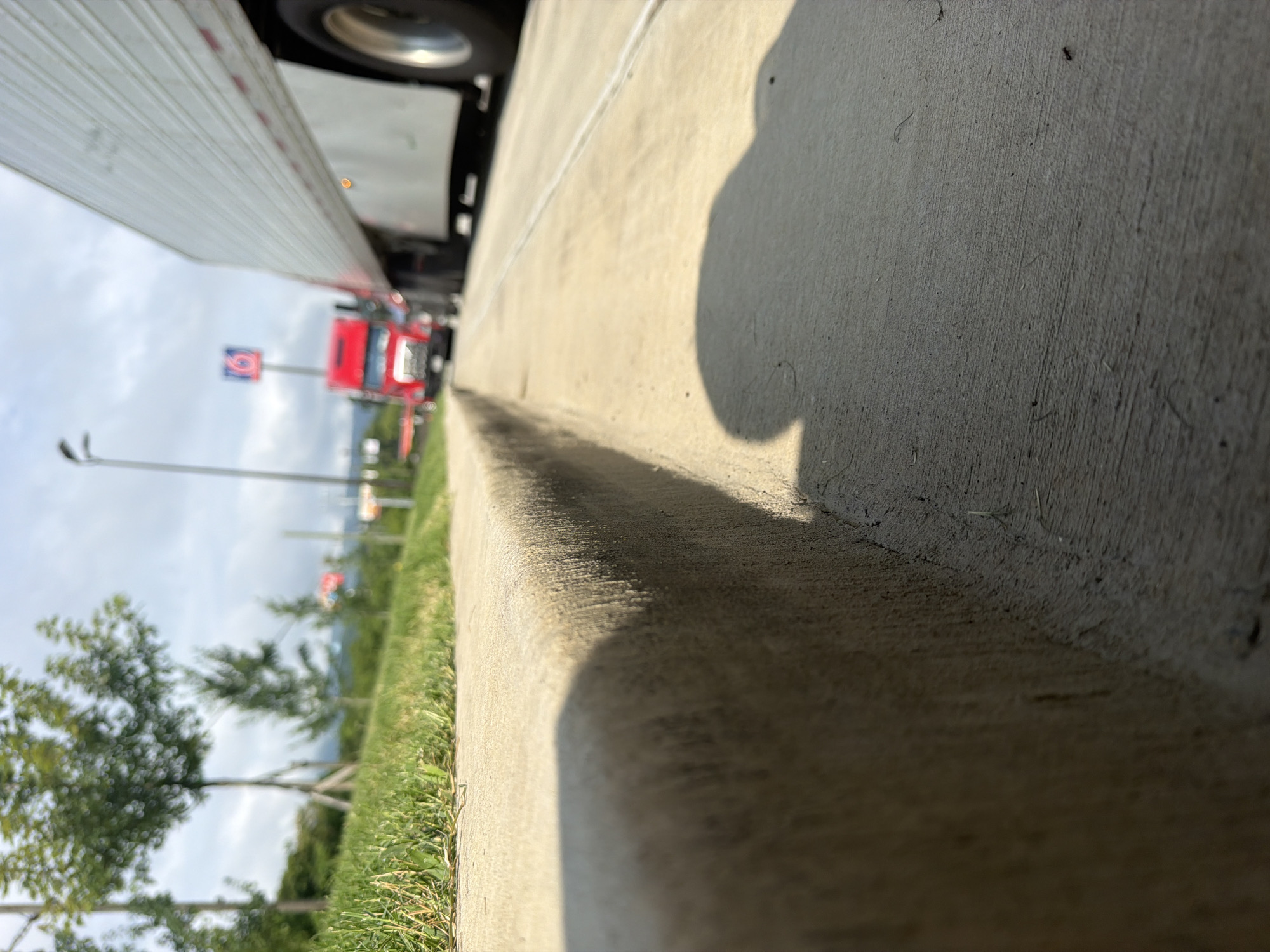 This image shows a low-angle view of a concrete curb on a sidewalk. In the background, there is a red semi-truck and a white semi-truck, possibly stopped at a traffic light. The red truck has "6" visible on a sign behind it. There are trees and grass to the left of the curb, and the sky is cloudy. The scene appears to be along a road in a city, with the distinct signage of a truck stop or gas station visible in the distance. The overall lighting suggests it is daytime. While the image is taken in Chattanooga, United States, specific landmarks or street names are not identifiable from the visual information provided.