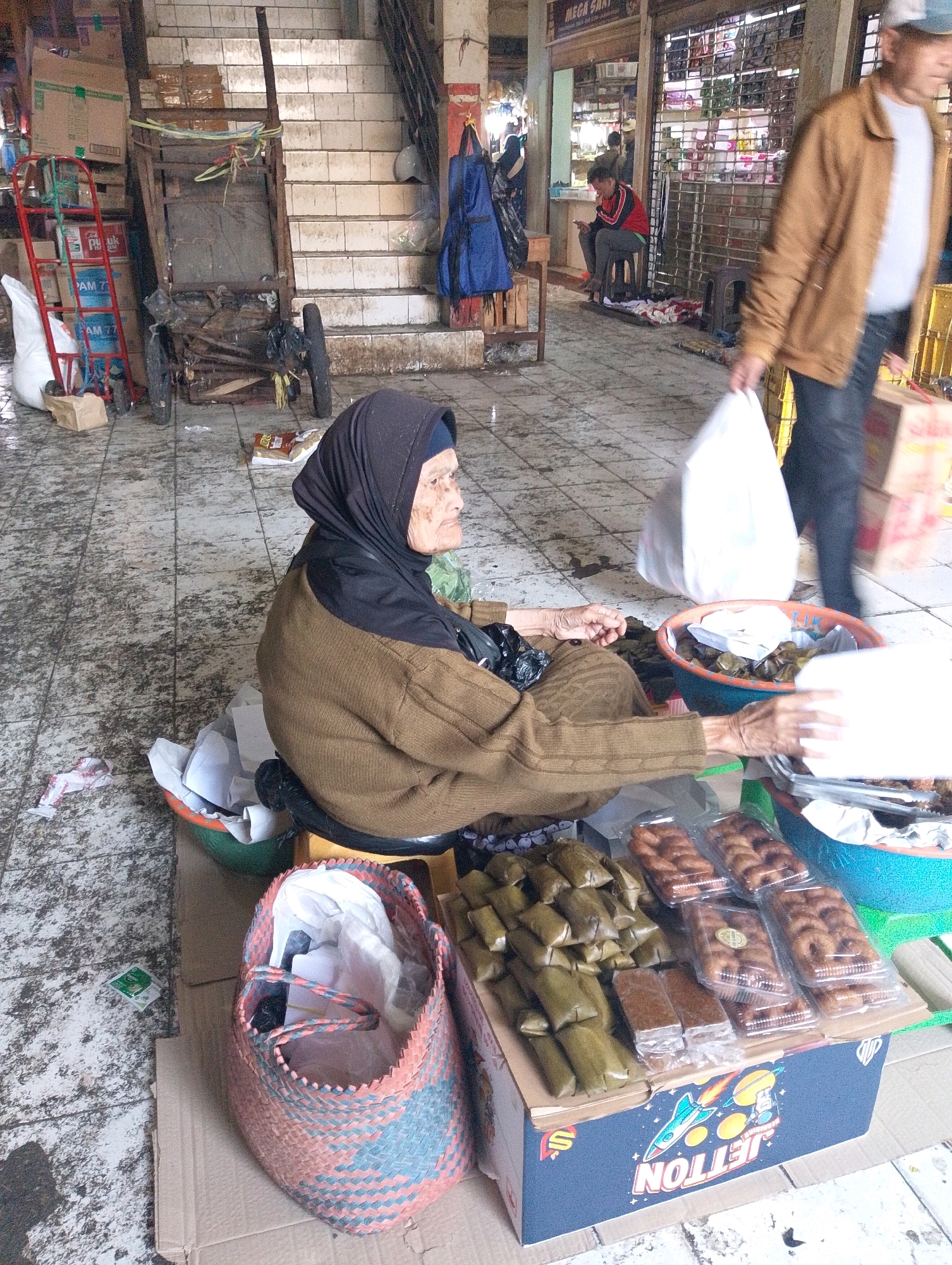 An elderly woman wearing a black hijab and a brown sweater sits on the floor of what appears to be a market in Lembang, Indonesia. She is looking to her right, with her hands positioned near a collection of food items. In front of her, a collection of packaged food, including wrapped rice cakes and fried pastries, is displayed on a blue cardboard box with "JETTON" branding. To her left, a woven basket filled with white plastic bags is visible. The ground is tiled and appears somewhat worn. In the background, a set of stairs leads up, and stalls with metal shutters are visible. A person in a tan jacket and dark pants walks by in the mid-ground, carrying a white plastic bag. Another person is seated in the background, looking at a phone. A metal cart with a red frame is positioned near the stairs, and various boxes and bags are scattered around the area.