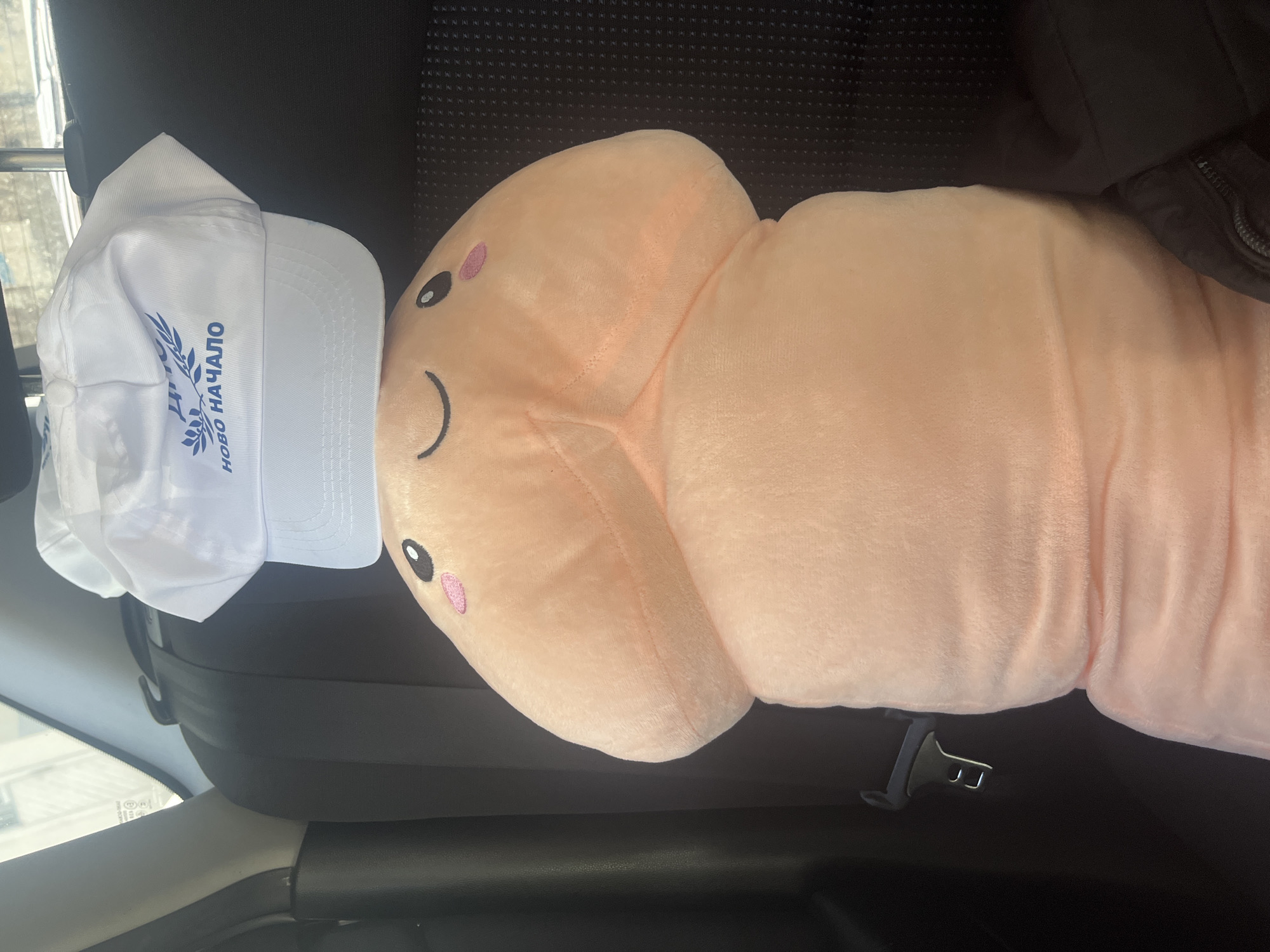 The image shows the interior of a car, specifically the passenger seat. A large, plush, light pink pillow resembling a penis is lying on the seat, secured by a seatbelt. On top of the pillow's "head" rests a white baseball cap with blue lettering and a wheat stalk logo, reading "НОВО НАЧАЛО" (Novo Nachalo). The interior of the car appears to be black. The location of Varna, Bulgaria, is not evident in the image.