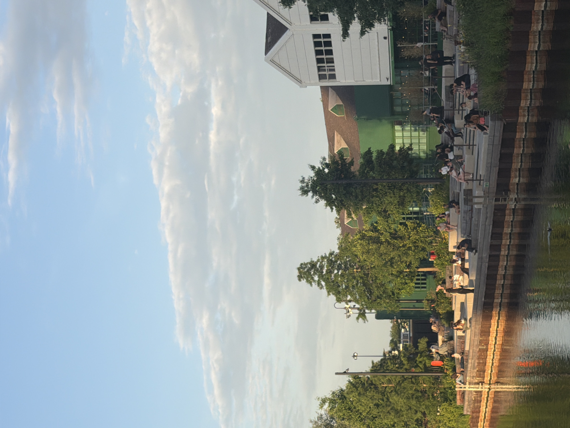 The image depicts a scene along a waterfront walkway, likely in London, UK, evidenced by the architectural style and the presence of a canal. A two-story white building with a dark roof, possibly a restaurant or community center, is visible along the walkway. A second building is green and appears to have people sitting along the edge of the walkway near the canal.
People are sitting and standing along the walkway, engaging in various activities such as socializing or simply enjoying the view. The walkway has tiered levels, perhaps steps. Trees and vegetation are interspersed along the walkway, providing shade.
The canal reflects the buildings and trees along its bank. The sky is light blue with some clouds.