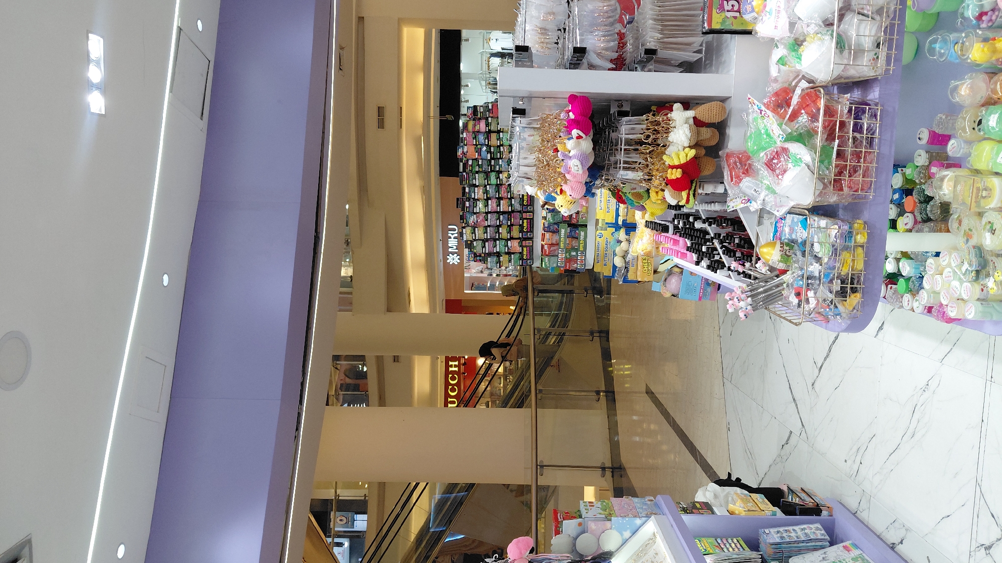 The image shows a brightly lit interior of a shopping mall, likely in Purwokerto, Indonesia. In the foreground, to the right, a display shelf is filled with various trinkets and novelty items, including keychains shaped like cartoon characters and small decorative items. Baskets on the shelf contain colorful packaged goods, possibly toys or candy. Further to the left, another display unit showcases more items, including stationery and packaged goods. In the background, escalators are visible, suggesting a multi-level mall. Two individuals are seen on the escalator, one wearing a hijab. The mall interior features white marble flooring with a patterned design, and cream-colored pillars. A large purple wall section is prominent in the upper part of the image, adorned with discreet white lighting strips. A store sign with the logo "MIKU" is visible, along with displays of products. The overall atmosphere is that of a commercial retail environment.