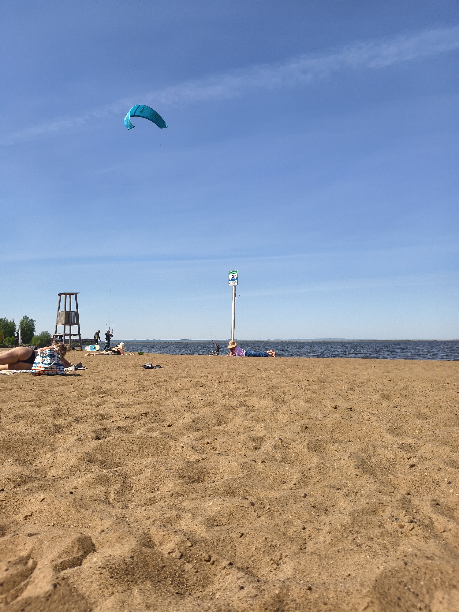 The image depicts a beach scene with a sandy foreground leading to a body of water, possibly a lake or sea. In the background, there is a clear blue sky with a single, blue kite flying high. Several people are scattered across the beach. To the left, a lifeguard tower is visible, and closer to the center, there is a signpost. Some individuals are lying down, possibly sunbathing, while others appear to be involved in water sports, potentially kite surfing given the presence of the kite. The location is Unknown, Unknown.