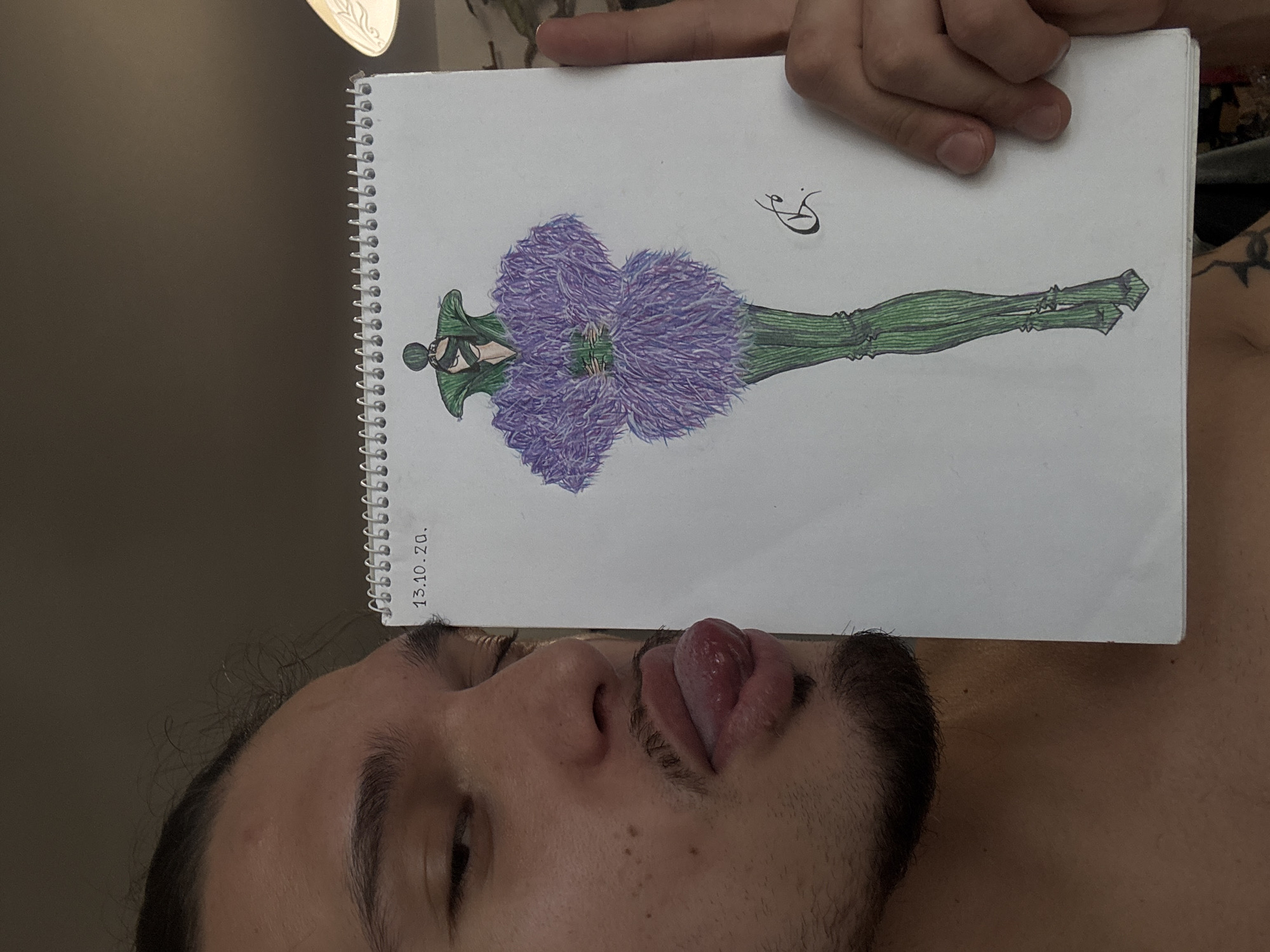 A man with dark hair tied back and a light beard is shown in a close-up, slightly angled shot. He is sticking his tongue out towards the camera. To his right, he holds a white spiral-bound sketchbook open, revealing a fashion illustration. The drawing depicts a full-body figure wearing an outfit with a green, high-collared top and a voluminous, feathered purple dress with green accents. The bottom half of the outfit consists of green, textured leggings and pointed shoes. A date, "13.10.20.", is written at the top left of the drawing, and a stylized signature is present on the right side of the illustration. The background is dark and out of focus, suggesting an indoor setting. No specific location in Sofia, Bulgaria is discernible from the image.