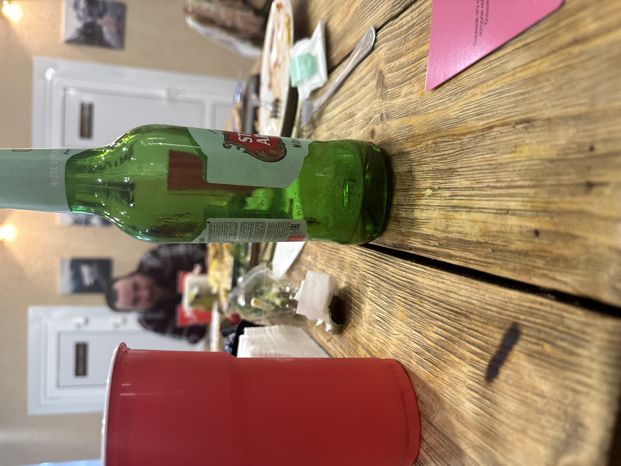 The image depicts a casual indoor setting, likely a gathering or meal, centered around a rustic wooden table. In the foreground, a prominent red plastic cup is visible. A green glass beer bottle, identified as Stella Artois, lies horizontally across the mid-ground of the table. Its labels feature "STELLA ARTOIS," "BIERE SUPER," "NON," and Cyrillic text including "КОРПОВАНОЕ ОБЪЕМ 0,44 Г" and "ЗЕЛ АЛКОГОЛЬНОЕ," suggesting its origin is consistent with the Chaikovsky, Russia location.
Other items on the table include a plate with remnants of food, a plastic fork, a small white packet, and stacks of paper napkins. A pink card is partially visible on the table, displaying fragmented Cyrillic text such as "Передай эту ка...", "которая м...", and "больше." In the blurred background, a man with dark hair or a beard is faintly visible, looking in the direction of the camera, indicating he is present at the event. Wall decorations, including framed pictures and a white door frame, are also discernible in the background. The scene suggests a relaxed atmosphere, likely taking place in the evening hours indoors.