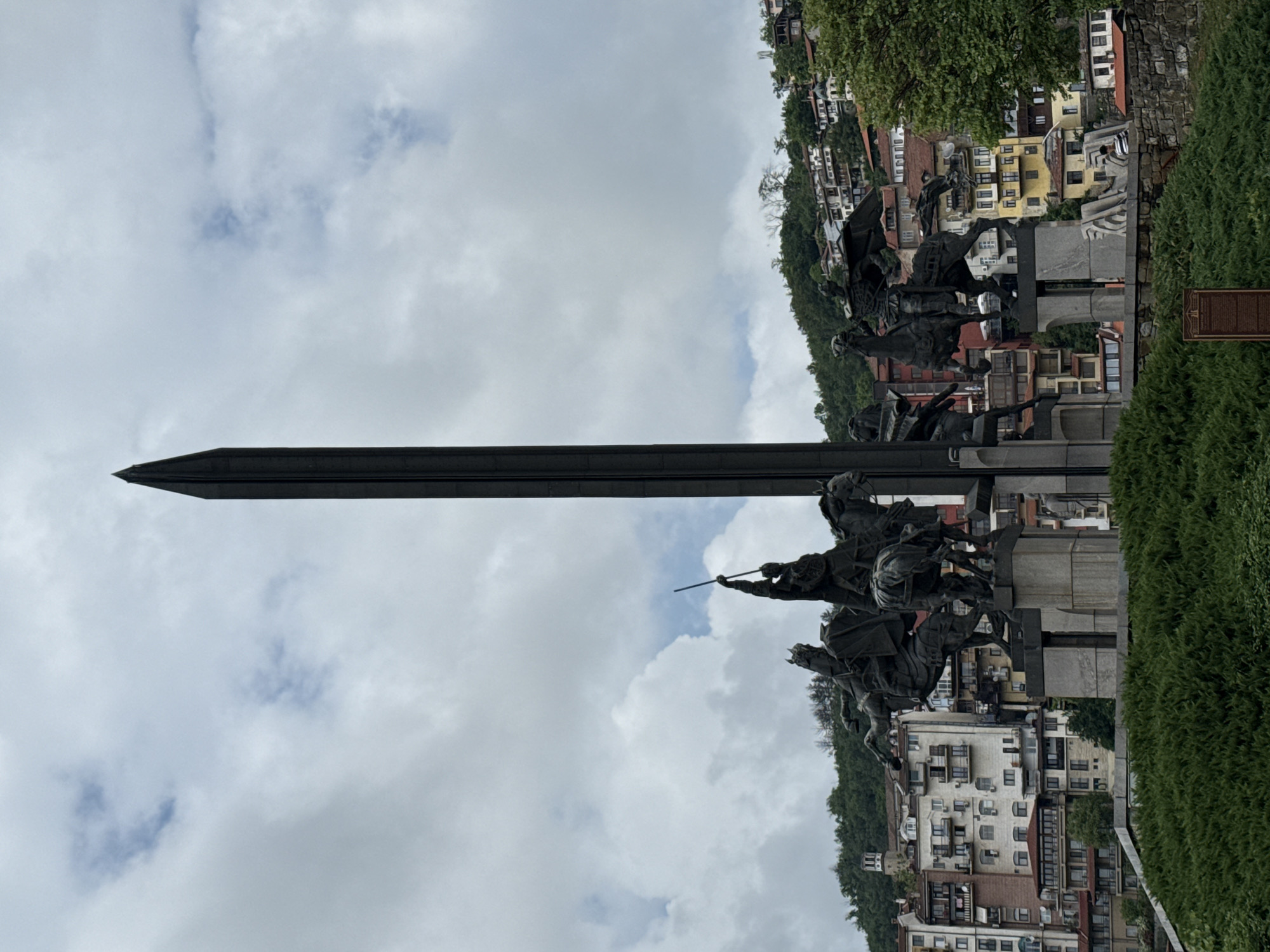 The image shows the Asenevtsi Monument in Veliko Tarnovo, Bulgaria. The monument depicts four kings on horseback in the foreground. A tall, dark sword-like structure rises behind them toward the sky. The city of Veliko Tarnovo, with its buildings and green trees, is visible in the background. The sky is overcast with white and gray clouds.