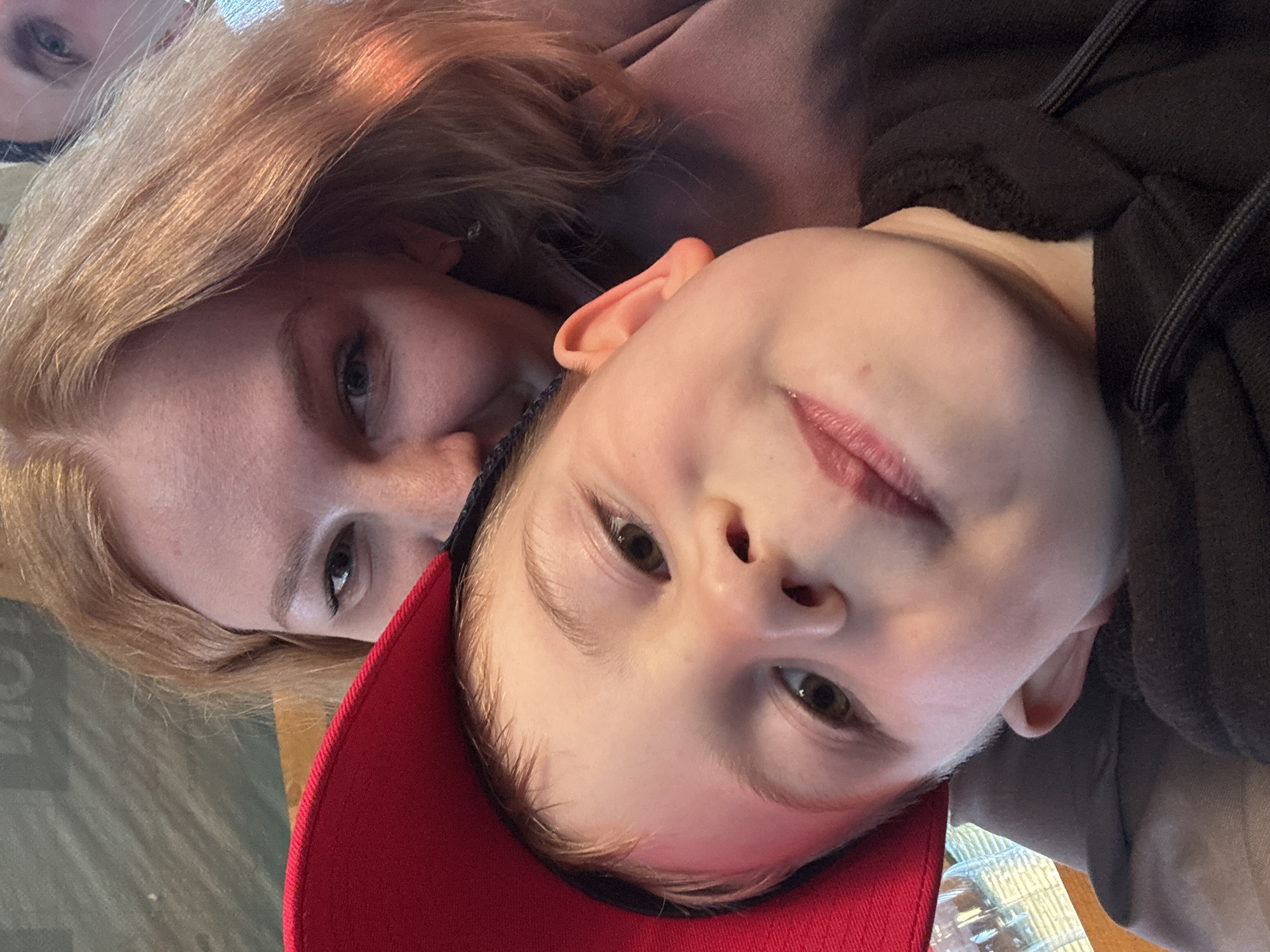 This image depicts an adult female and a young male child in an indoor setting, specified as the city of Rozhdestveno, Russia.
The adult female is positioned in the upper right quadrant, partially obscured by the child. She has light-colored, wavy hair, parted on her left side. Her visible left eye features dark eyeliner. A subtle upward curve is present at her mouth, consistent with a faint smile. A small, reflective earring is visible in her right ear. She wears a garment of a muted reddish-brown or pinkish hue.
The young male child is in the foreground, angled towards the viewer with his head tilted. He wears a red baseball cap, with the brim positioned towards the viewer's left. His hair is light brown, visible beneath the cap. His eyes are dark brown and directed at the camera. His lips are slightly parted in a small smile or smirk. His left ear is visible. He is dressed in a black hooded sweatshirt with visible drawstrings.
The background is significantly out of focus. To the upper left, a blurry green-grey surface with faint, reversed text is discernible. In the upper right corner, a partial, blurred reflection of another individual's eye and forehead is present. A blurry, reflective object, possibly a glass or bottle, is partially visible in the lower left. The overall lighting is warm, indicating an interior environment.