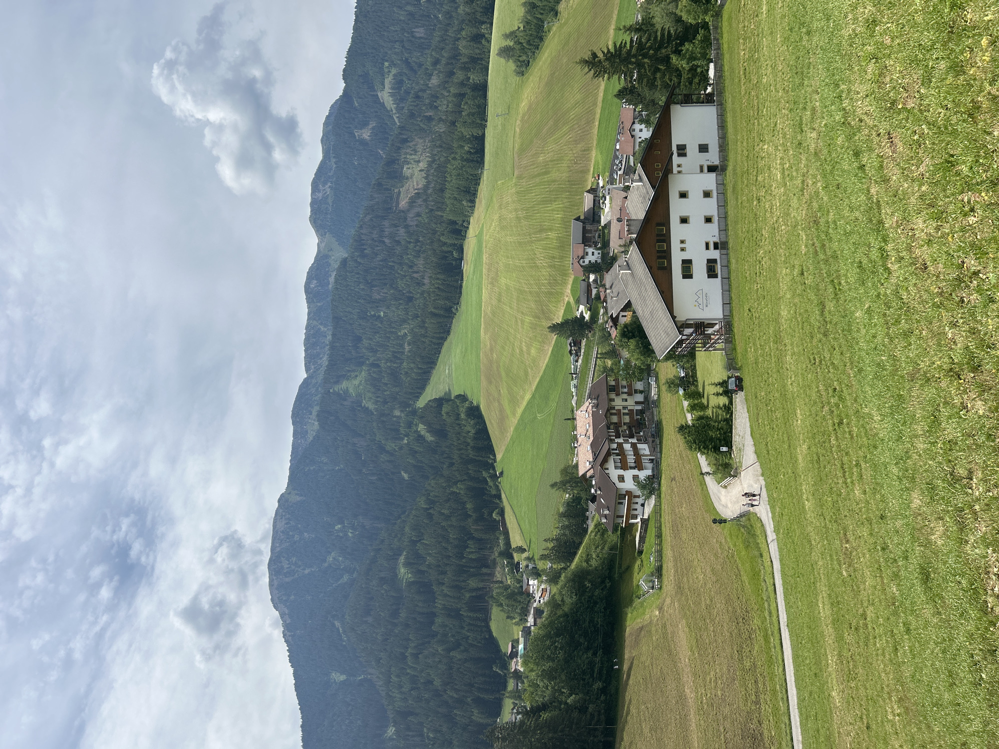 The image shows a scenic view of a village nestled in a valley, surrounded by lush green hills and a majestic mountain range in the background. The sky is partly cloudy, with dramatic clouds adding depth to the scene.
In the foreground, a vibrant green meadow slopes down towards a winding road. A few people are walking along this road, suggesting a leisurely stroll or hike.
Further into the mid-ground, a cluster of buildings forms the heart of the village. These buildings, primarily residential or small hotels, feature white facades with dark tiled roofs, characteristic of Alpine architecture. One building has a sign that reads "Mira Odlle" with a mountain logo.
Beyond the village, the landscape transitions into rolling green hills that are meticulously cultivated, with visible patterns from farming or mowing. These hills are interspersed with patches of dark green coniferous forests.
The background is dominated by a towering mountain range. The lower slopes are densely forested, while the upper reaches are rocky and rugged. A ski slope or cleared path is visible on one of the mountainsides, indicating potential recreational activities. The overall impression is one of tranquility and natural beauty, with human settlement harmoniously integrated into the mountainous environment.