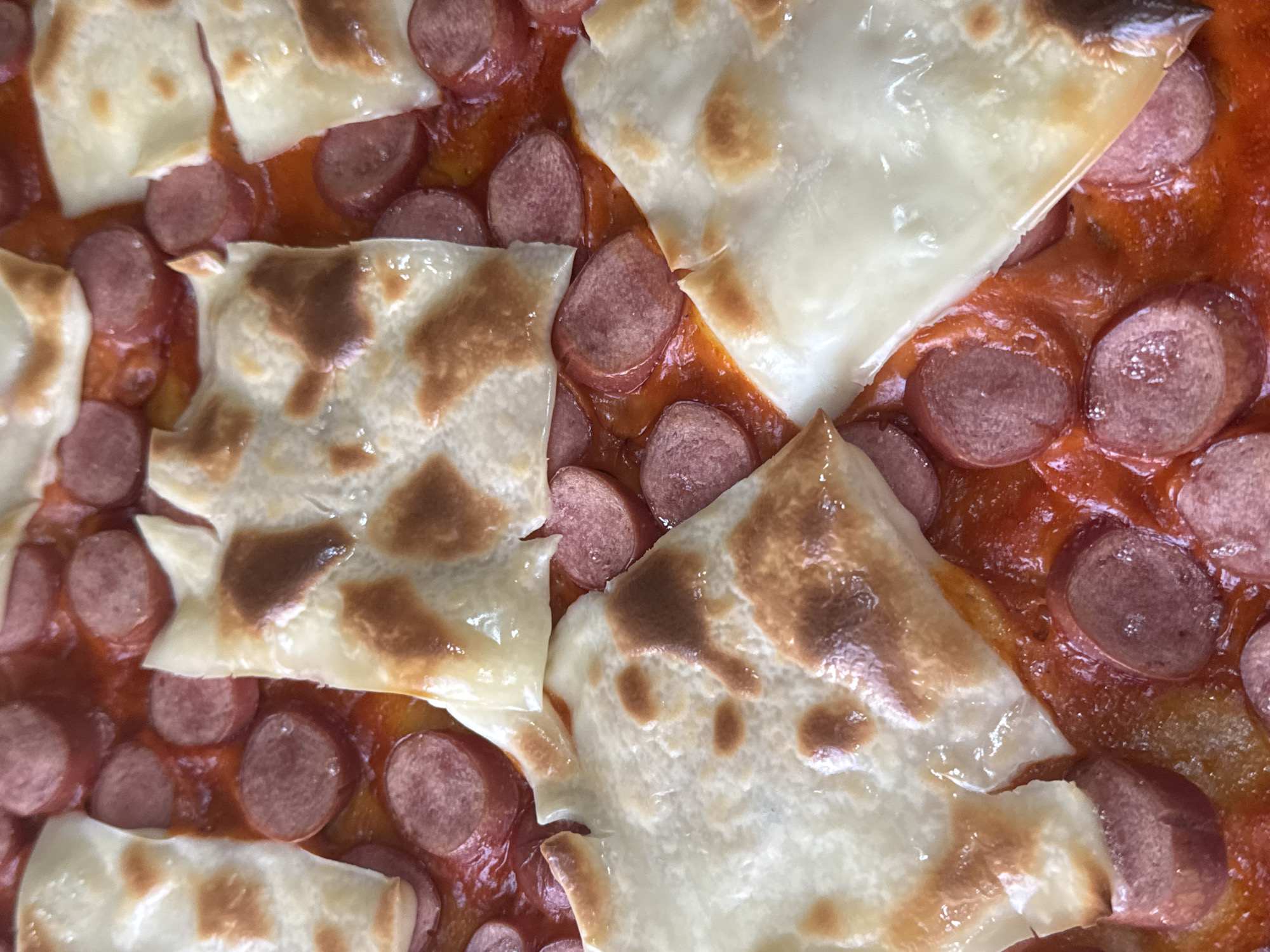 This is a close-up, top-down shot of a cooked food dish. The primary subjects are numerous circular slices of reddish-brown sausages or hot dogs, which are interspersed throughout a vibrant red, tomato-based sauce. Layered on top of the sauce and sausages are several large, irregularly shaped pieces of a light-colored, thin substance that appears to be melted cheese or possibly pasta sheets, such as lasagna noodles. These light-colored pieces exhibit varying degrees of browning, ranging from golden to dark brown, suggesting they have been baked or grilled. The sauce itself looks somewhat viscous and glossy, with small bubbles indicating its heated state. The food appears fully cooked and ready to serve. No activity is taking place, only the finished product is shown. There are no people visible, and no specific location or time of day can be determined from the image. No text is visible.
