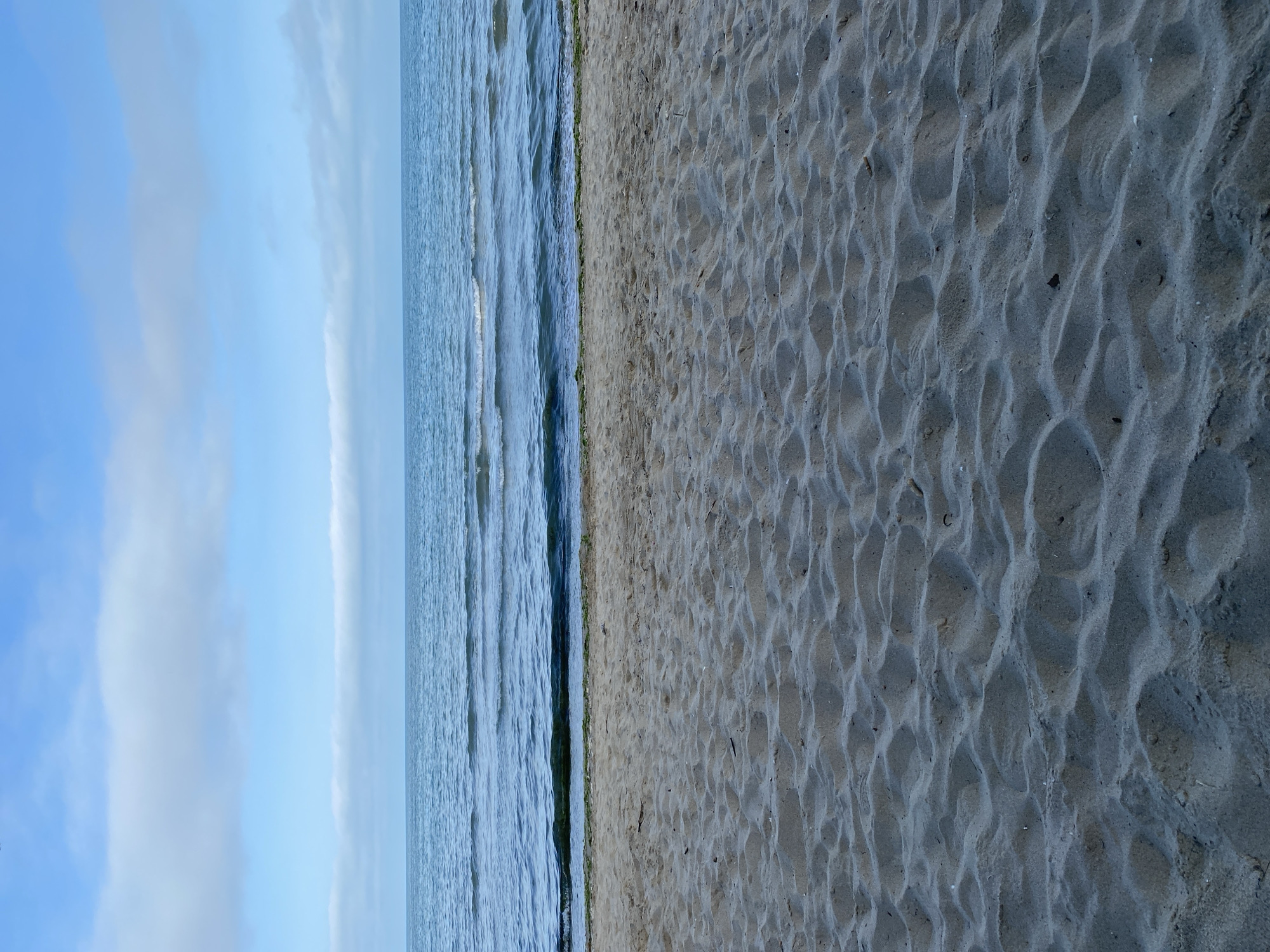 The image depicts a beach scene, divided into three main sections: sky, water, and sand. The sky takes up the left side of the image and shows blue skies. The water is in the center, with gentle waves visible. The sand occupies the right side of the image, displaying textured patterns. Based on the presence of these elements and context clues, this image is likely taken at a beach in Varna, Bulgaria. However, without more specific information (e.g., landmarks, beach names), pinpointing the exact location within Varna is difficult.