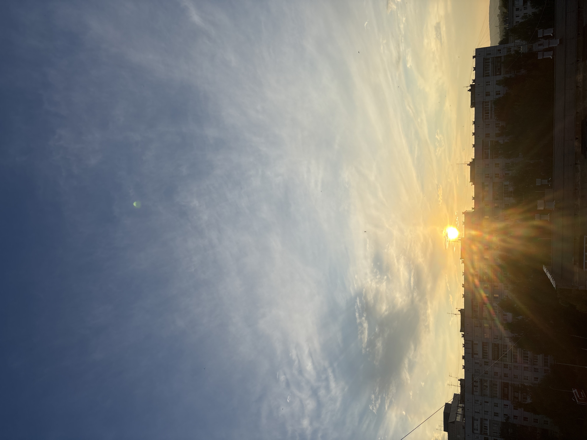 The image depicts a sunset over an apartment building in Chaikovsky, Russia. The sun, a bright orange orb, is positioned just above the horizon, casting long rays of light that create a starburst effect. The sky is a gradient of blue at the top, transitioning to yellow and orange near the horizon, with wispy white clouds scattered throughout. The apartment building is a long, multi-story structure with many windows and balconies. Trees are visible in front of the building, silhouetted against the bright sky. The overall scene is one of a serene urban landscape at dusk.