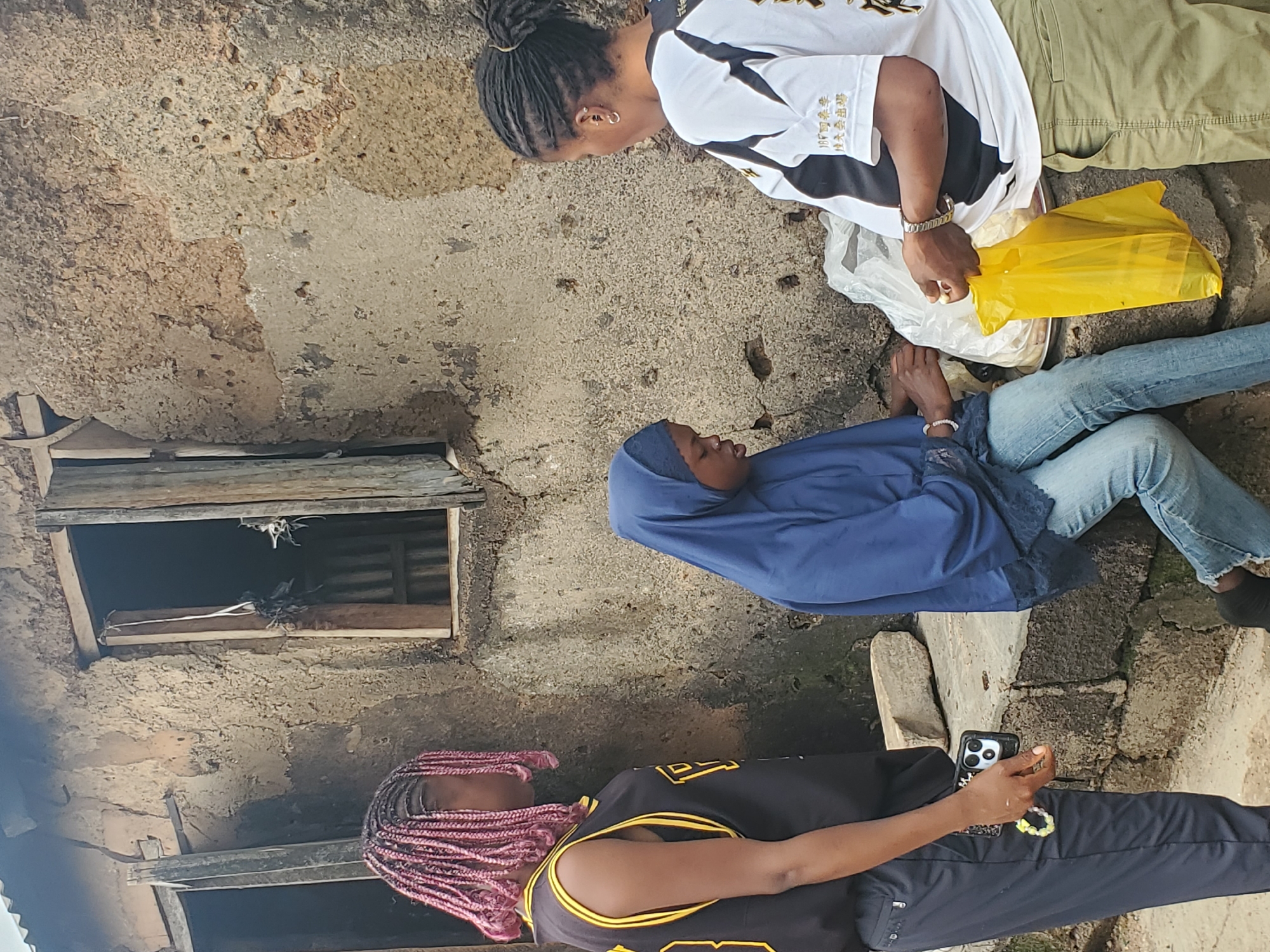 The image shows four people in what appears to be an outdoor setting with rough, weathered walls. The people are positioned in different ways, with two standing and two seated or reclining.
In the background, a person wearing a white t-shirt with black accents and khaki pants is standing and holding a yellow plastic bag. To their left, another person is leaning down, looking towards the ground, wearing a white t-shirt with black trim and what appears to be writing in a foreign language.
In the foreground, a person is seated, wearing a blue hijab and jeans. Next to them, another person with pink braided hair is lying down, wearing a black tank top with yellow trim. This person is holding a smartphone.
The scene has a raw, unfinished quality. The walls are textured and show signs of age and wear. There are two openings in the wall, resembling windows or alcoves, one of which contains some debris. The ground is rough and uneven. The lighting suggests it is daytime. The overall impression is one of a casual gathering in a rustic environment.