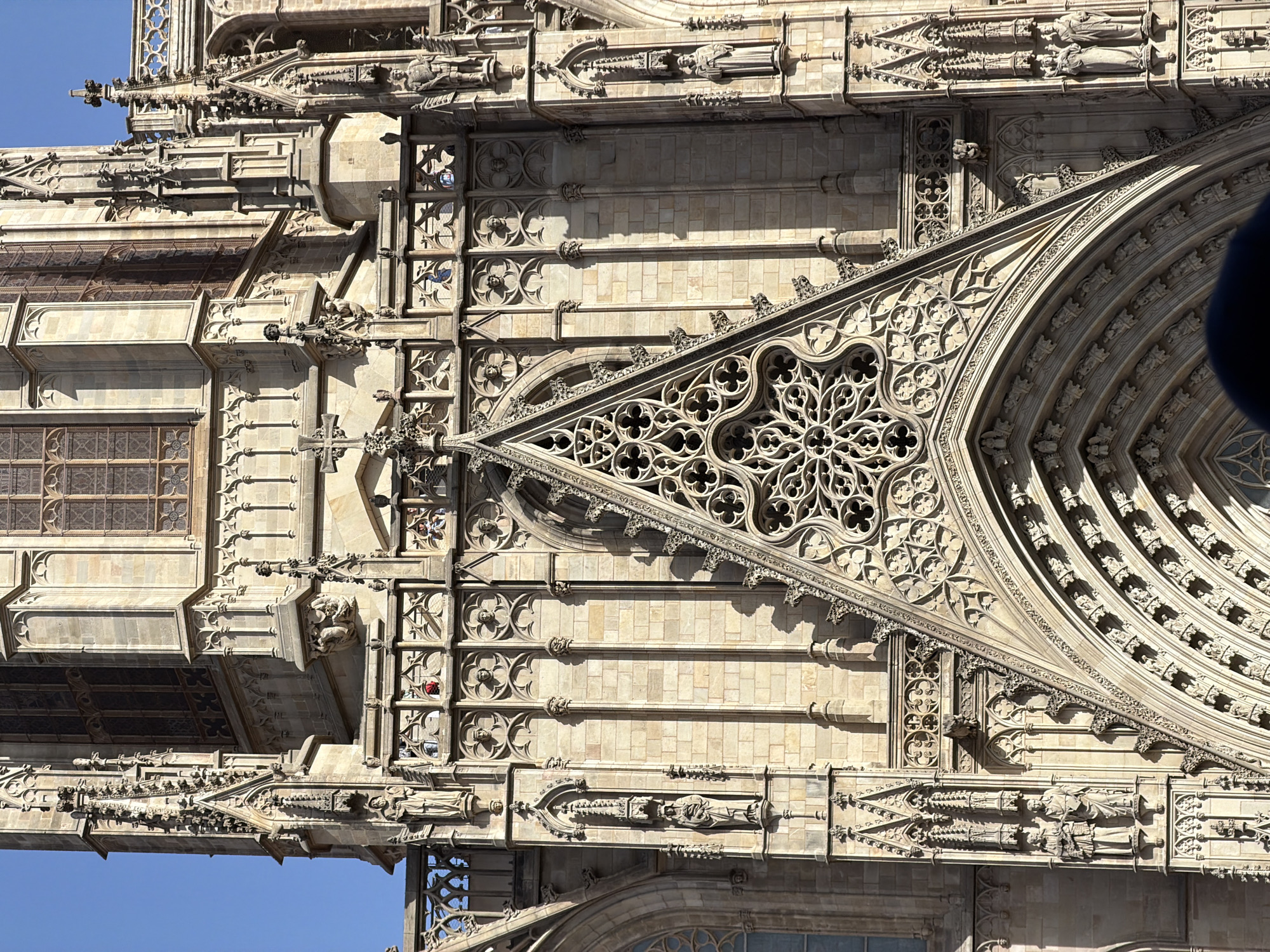 The image shows a detailed view of the facade of Barcelona Cathedral, a historic Gothic cathedral located in the Gothic Quarter of Barcelona, Spain. The perspective is from below, looking up at the intricate stone carvings and architectural elements. The stone is a warm, sandy color, and the bright sunlight creates strong shadows that highlight the detailed relief work.
The main focus is on a large, triangular pediment above an arched entrance, adorned with a complex rose window featuring geometric patterns of interlacing stone tracery. Below the rose window, the arched entrance is framed by multiple layers of decorative moldings and sculptural figures.
On either side of the central pediment, there are vertical sections of the facade that extend upwards. These sections contain niches with sculpted figures of saints and religious figures, as well as ornate pinnacles and buttresses. There are also horizontal bands of decorative stonework and smaller windows with stained glass.
In the upper portion of the image, parts of the cathedral's tower and upper windows are visible against a clear blue sky. Some small figures of people can be seen on ledges and balconies high up on the building, appearing as tiny silhouettes against the stonework. Their presence adds a sense of scale to the grandeur of the architecture. The overall impression is one of immense detail, craftsmanship, and the imposing presence of a significant religious structure.