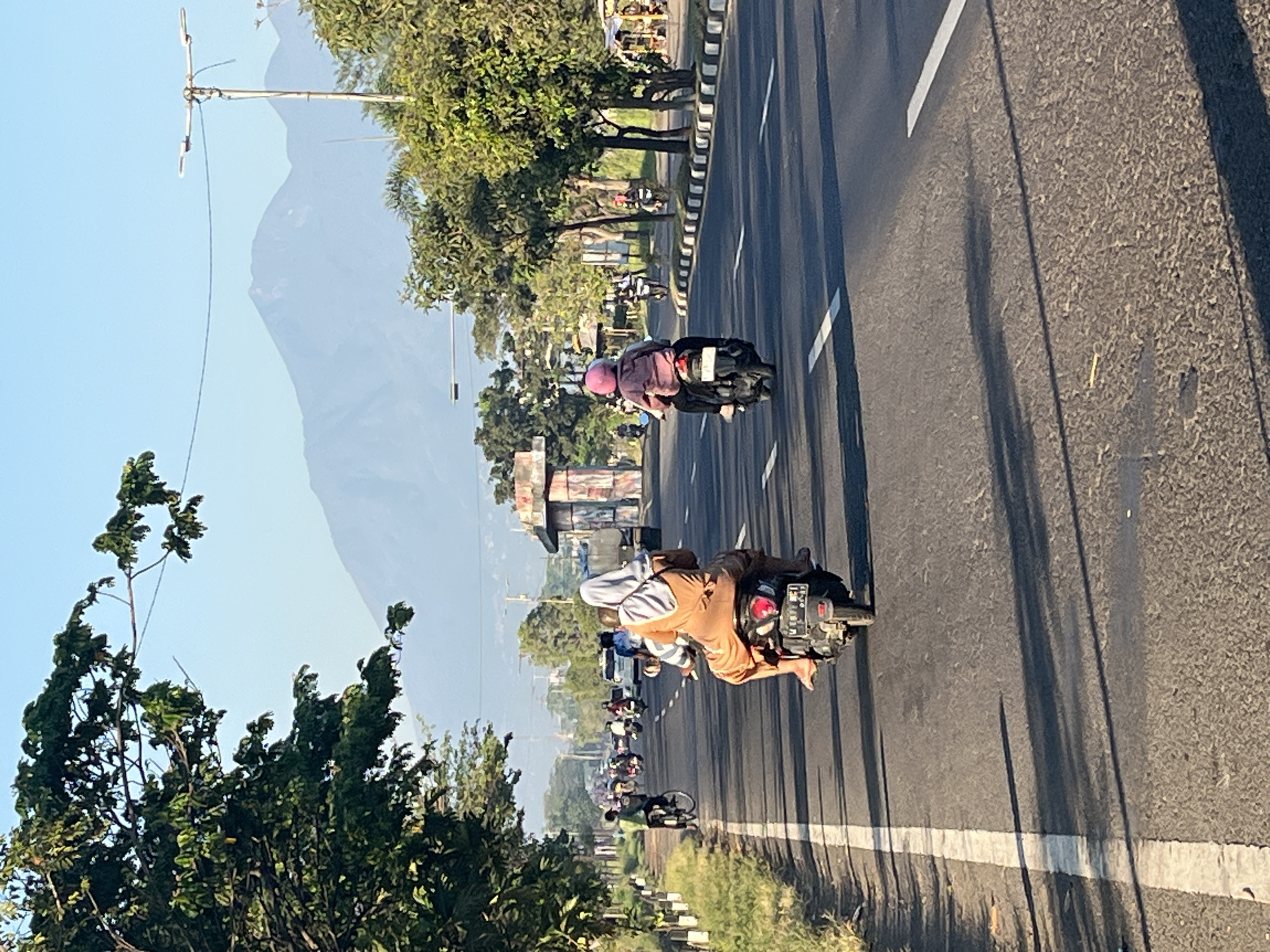 The image depicts a street scene in Siring, Indonesia, with a mountainous landscape in the background. Several motorcycles are traveling on the asphalt road. In the foreground, a motorcycle carries two people, a driver and a passenger. The passenger, wearing a beige hijab and a long beige dress, is seated behind the driver. The driver, with their legs visible, is operating the motorcycle. Further down the road, another motorcycle is visible with a person wearing a pink helmet and jacket. The road is lined with trees and a white line on the left side. In the distance, a large mountain range is visible under a clear blue sky. The overall scene suggests a typical day in a semi-urban or suburban area of Indonesia.