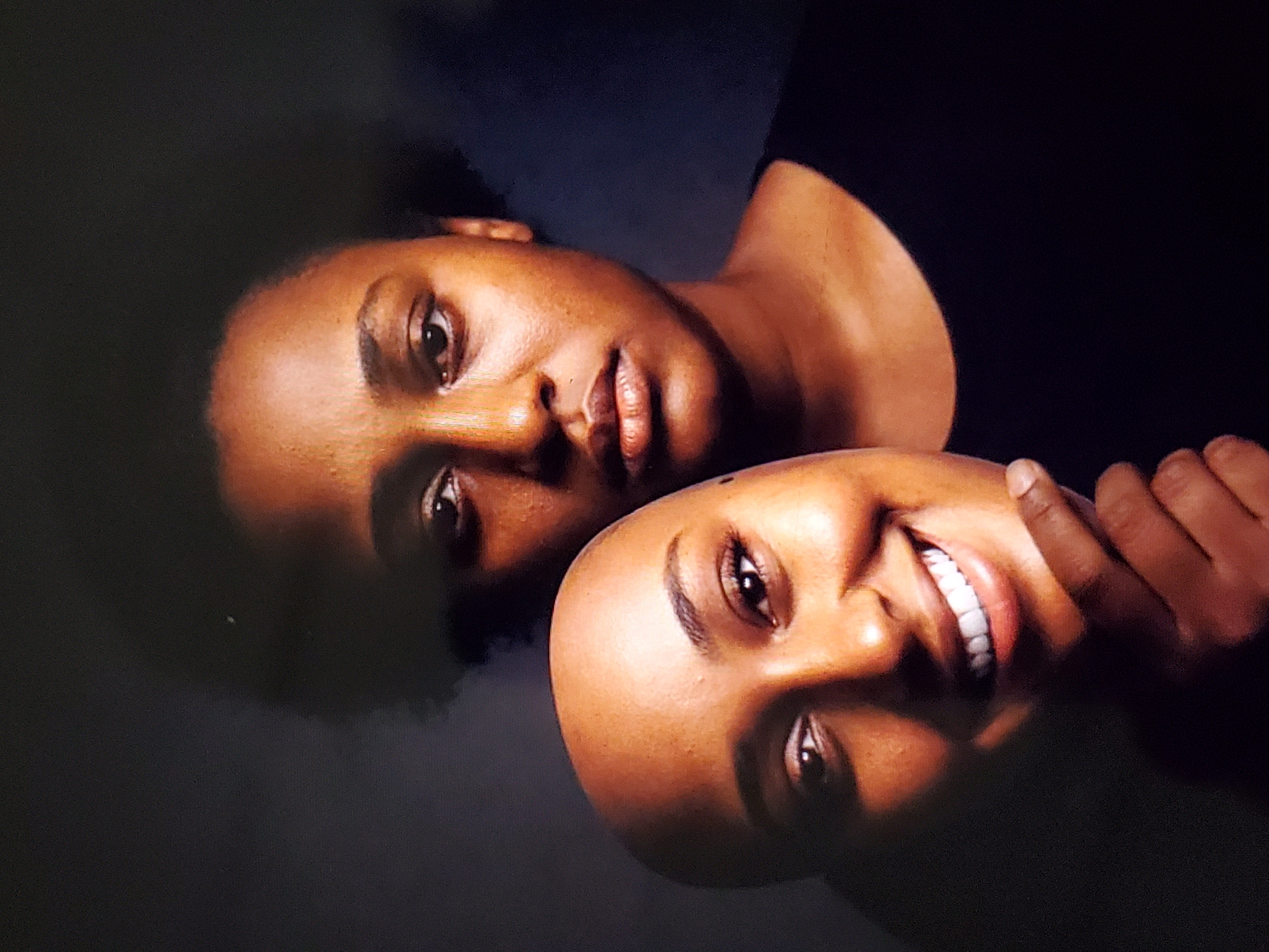 A medium shot shows two Black women in front of a dark background. The woman in the foreground is holding a mask that covers her face, revealing only her chin and a portion of her neck. The mask depicts a smiling woman's face. The woman in the background is looking directly at the camera, with her face partially obscured by the mask held in front of it. She has dark hair and is wearing a black t-shirt. The lighting is dramatic, casting shadows and highlighting the faces.